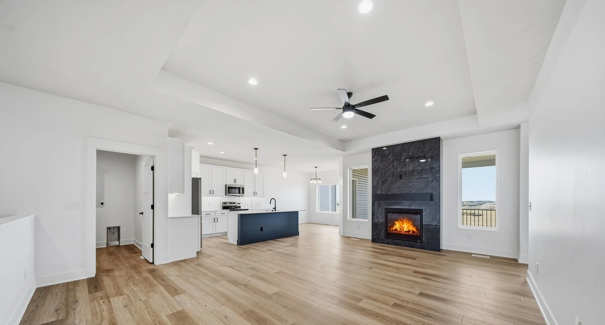Bright open living room with a modern black ceiling fan, a fireplace with a black marble surround, white walls, hardwood floors, and large windows showing a landscape view.