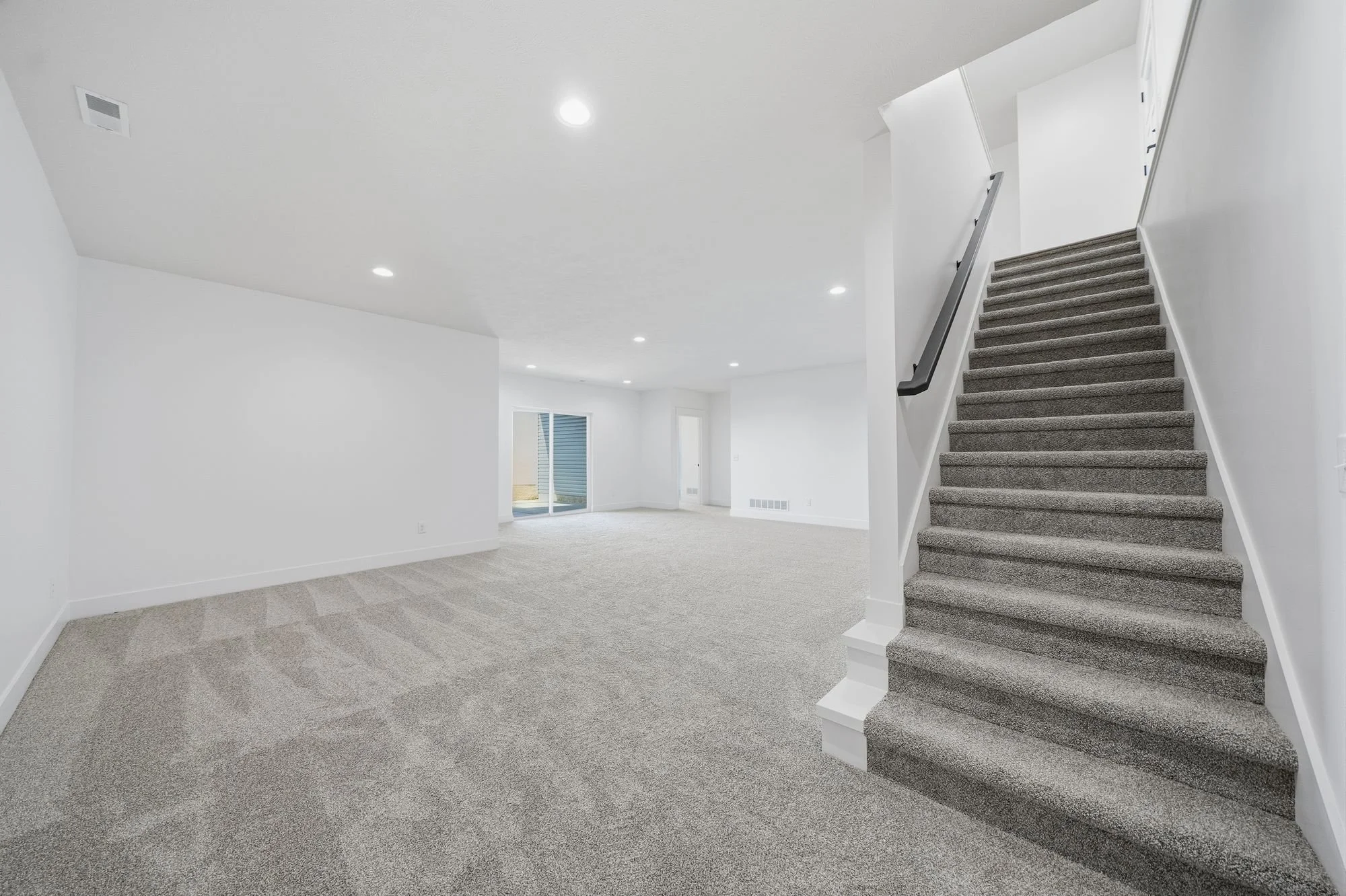 Empty living room with gray carpet and white walls, staircase with gray carpet and black handrail, sliding glass door leading outside, and recessed ceiling lights.