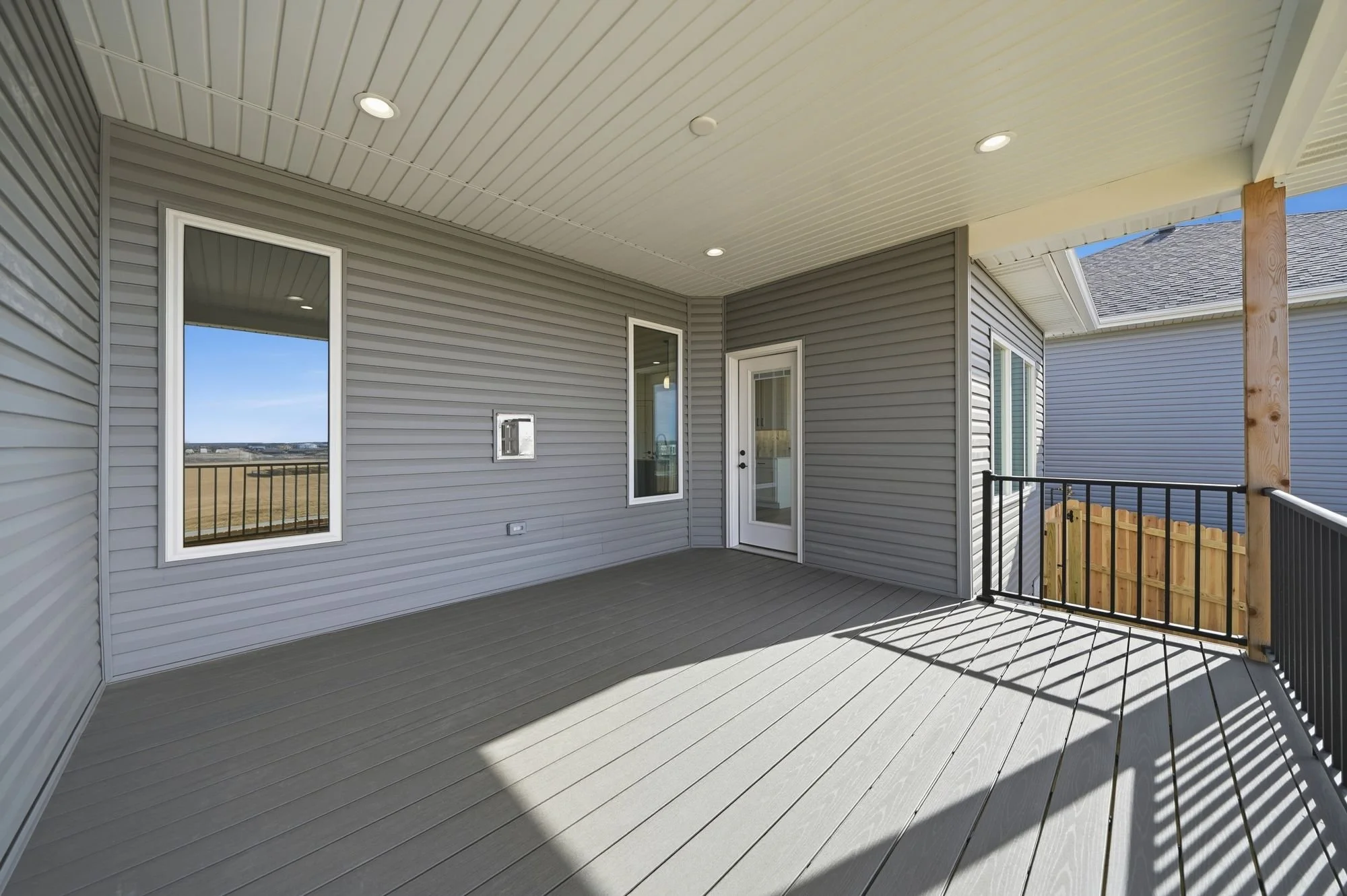 A covered balcony or porch with gray siding, two windows, a glass door, a black metal railing, and a wooden support post, with a view of neighboring houses and a blue sky.
