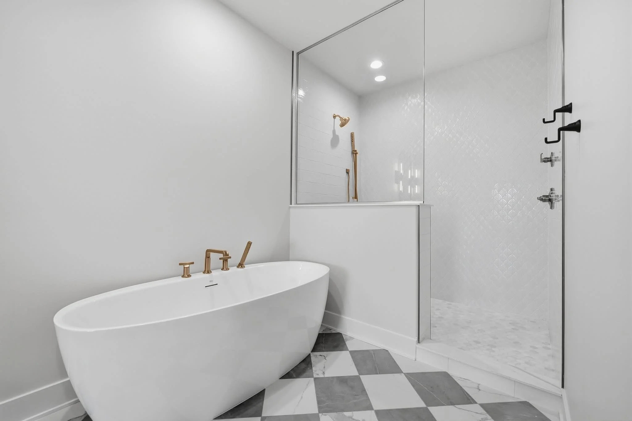 A modern bathroom featuring a white freestanding bathtub with gold fixtures, a glass-enclosed shower with a textured white tile wall, black hooks on the door, and a marble checkered floor.
