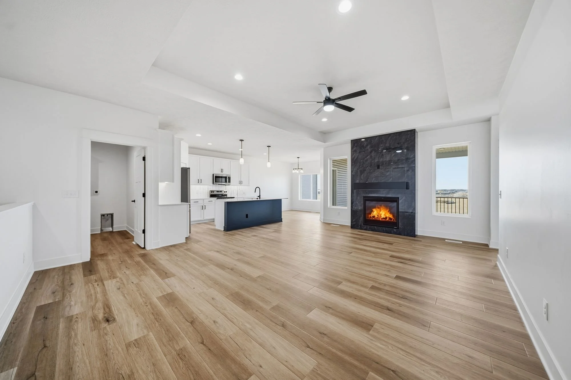Modern open-concept living room with wooden flooring, a black ceiling fan, a fireplace with a black marble surround, white walls, and large windows showing a view outside.