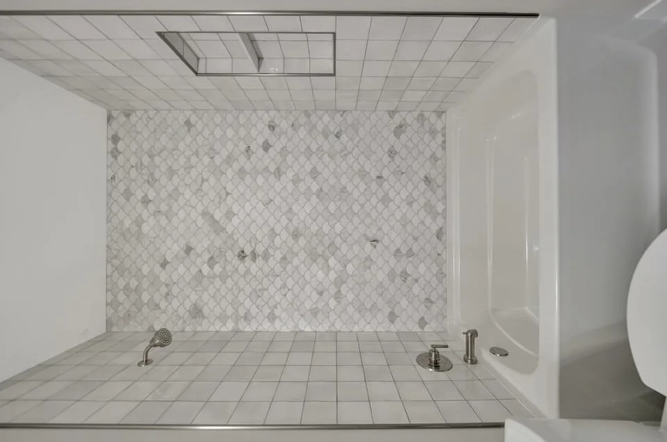 Bathroom with white square tiles, a shower with a handheld showerhead, a bathtub, and a wall with small marble tiles.