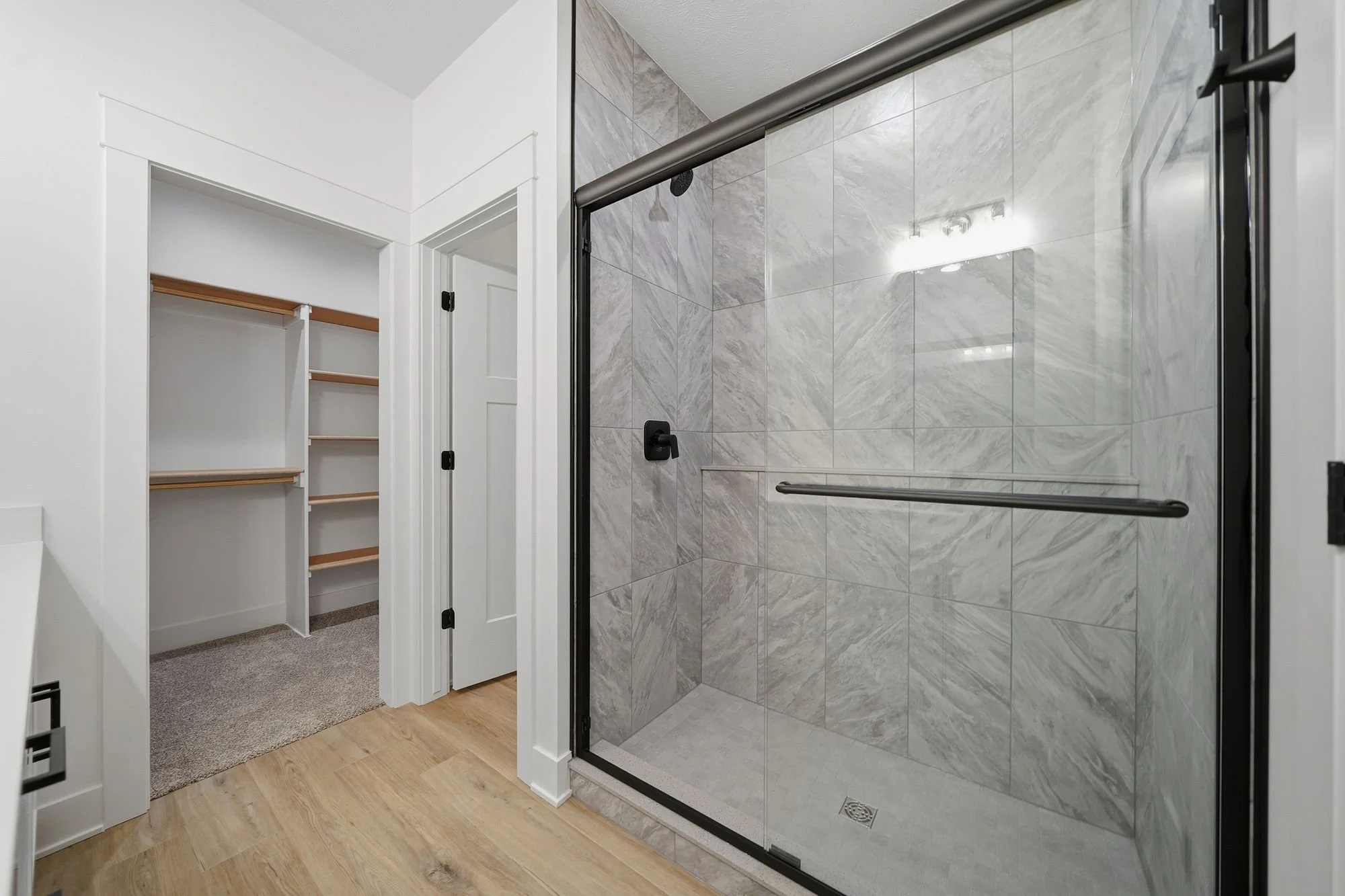 Bathroom with large walk-in shower with glass door, gray marble tiles, black fixtures, and a grab bar. Adjacent to the shower is a closet with white shelves and wooden rods, with beige carpet and wood flooring visible.
