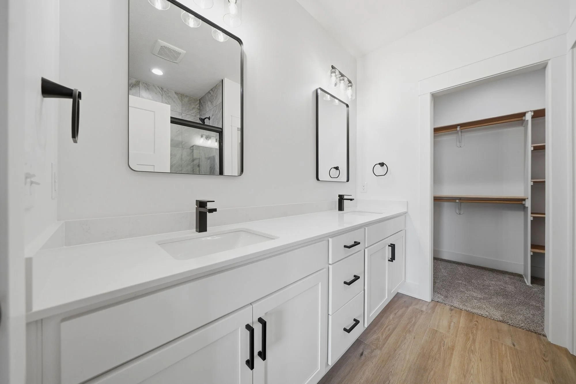 Modern white bathroom vanity with double sinks, black fixtures, and a large mirror, with a walk-in closet area visible in the background.