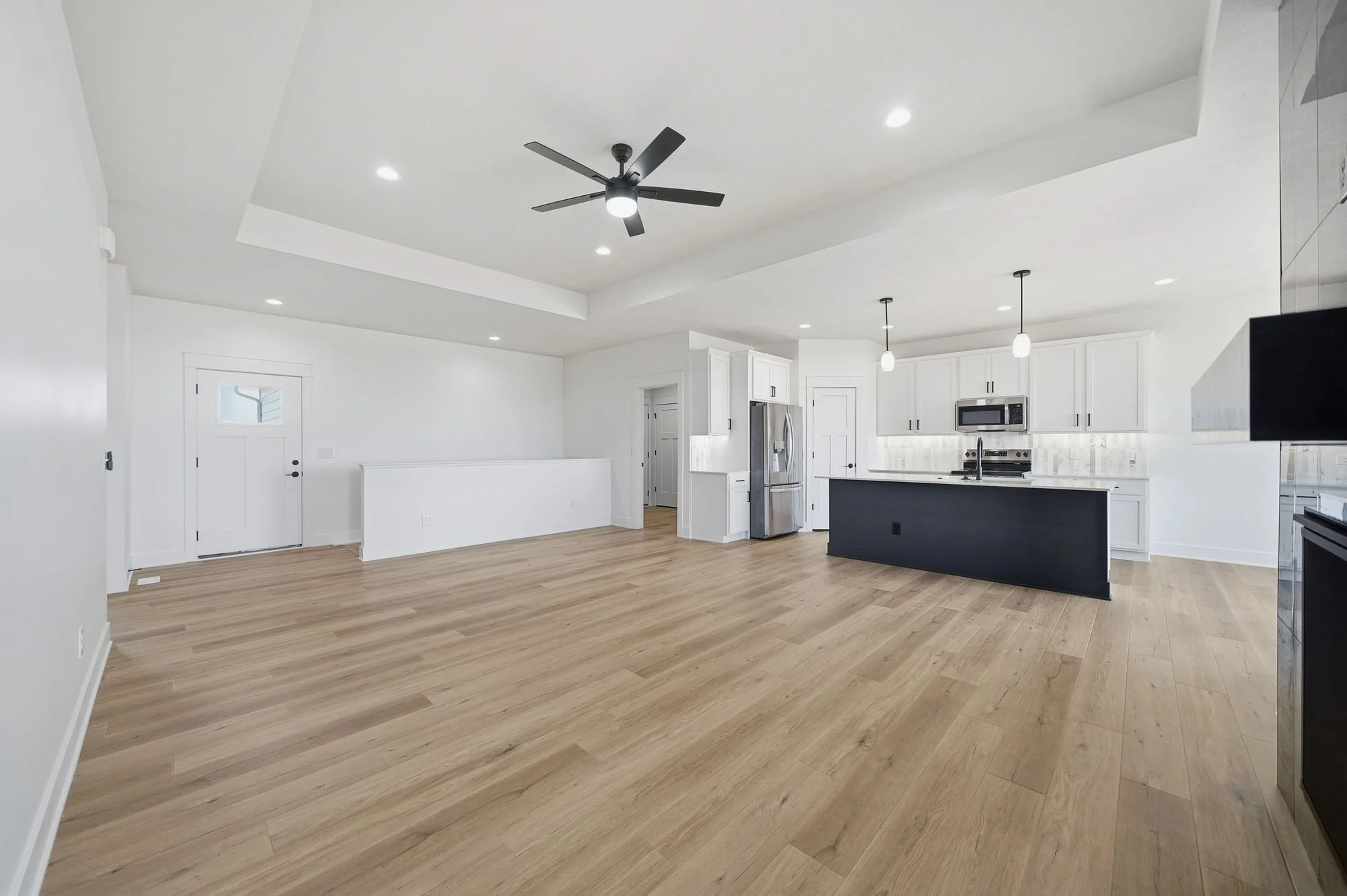 Modern open-concept kitchen with white cabinets, stainless steel appliances, and a black kitchen island. Light wood flooring and a ceiling fan with lights. Recessed lighting and hanging pendant lights.