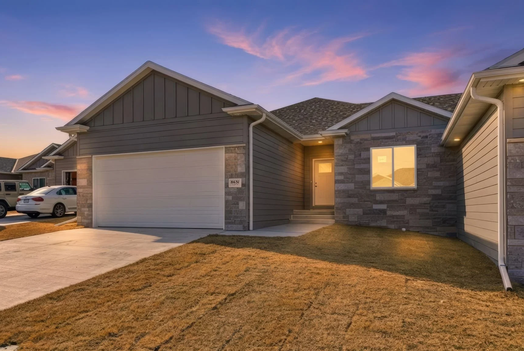 Front exterior of a modern house during sunset, featuring a stone and siding facade, a white garage door, a front door with steps, and parked cars in the driveway.