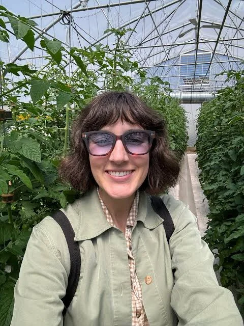 Woman wearing sunglasses smiling inside a greenhouse with green plants and vine trellises.