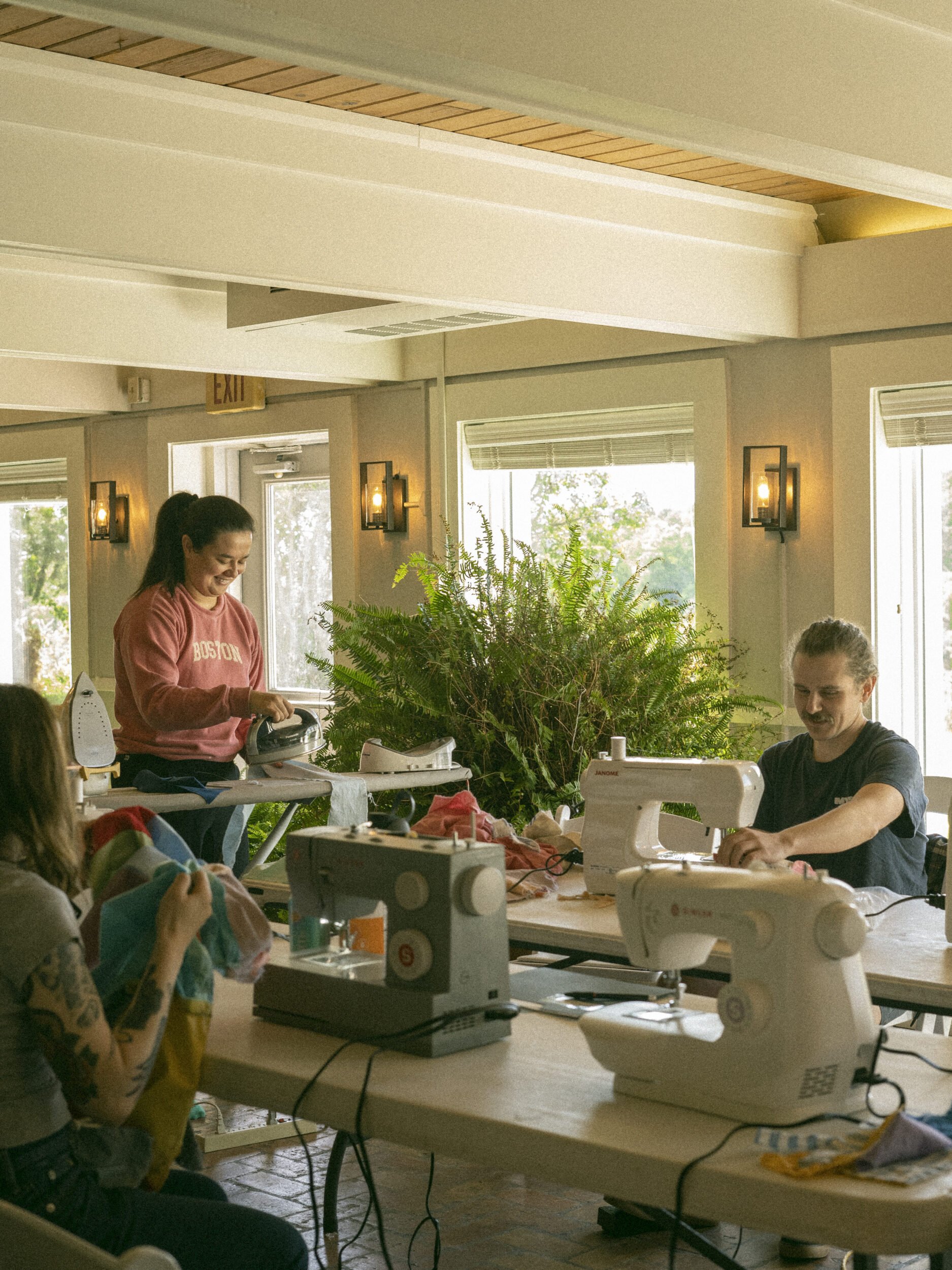 People sewing with multiple sewing machines at a table, bright natural light from windows, indoor space with plants and wall lamps.