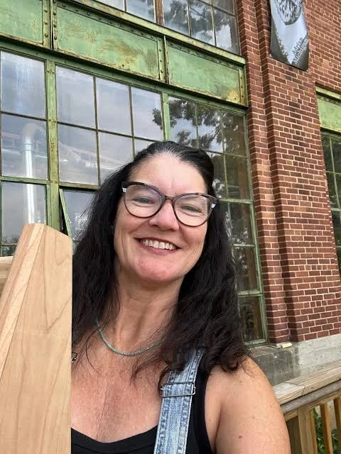 Woman with glasses smiling outdoors, standing in front of a large, old green metal and glass window on a brick building.