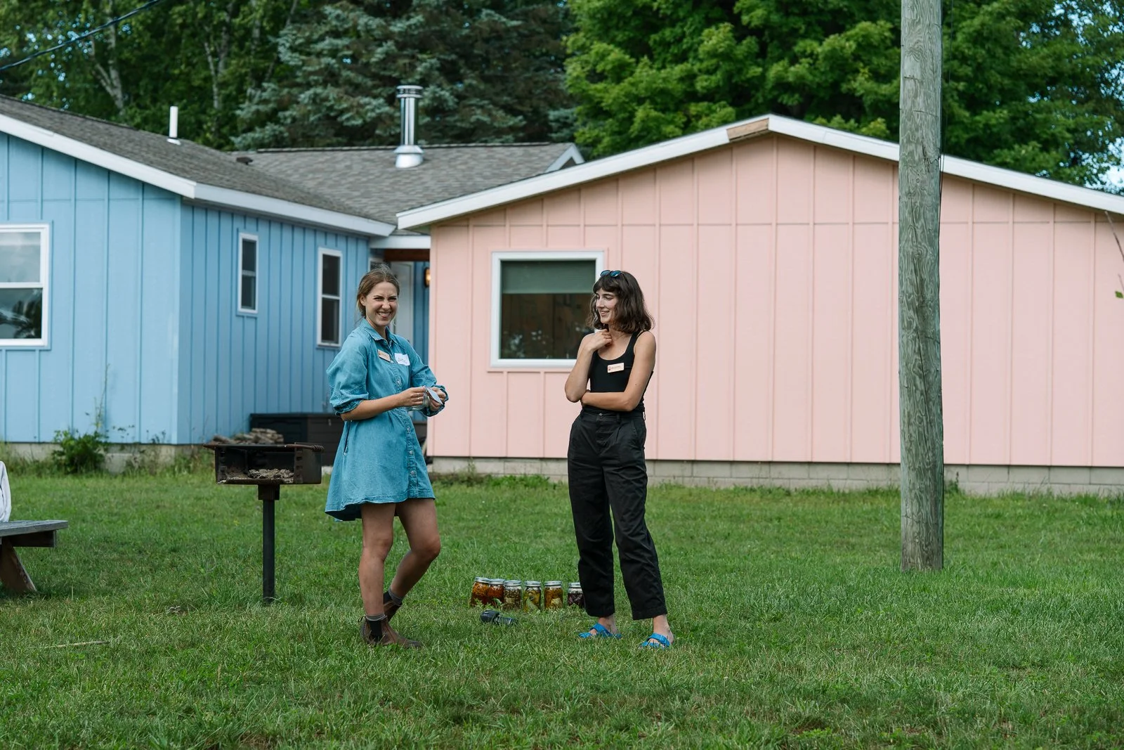 Two women standing on a grassy yard having a conversation, with a pink and blue house in the background. One woman is wearing a blue dress and the other in black pants and a black top, with jars of food on the ground between them.