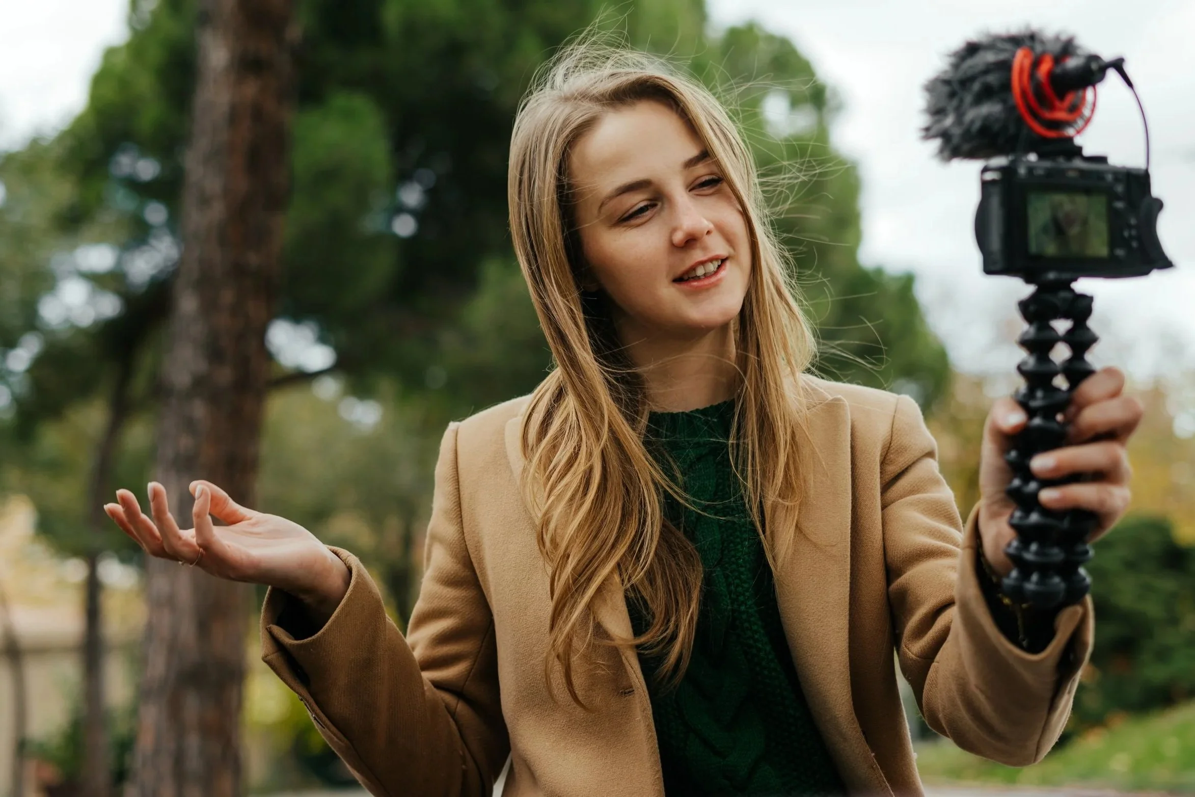 Junge Frau filmt mit einer Kamera im Freien, umgeben von Bäumen, sie spricht und gestikuliert. Sie trägt eine beige Jacke und einen grünen Pullover.