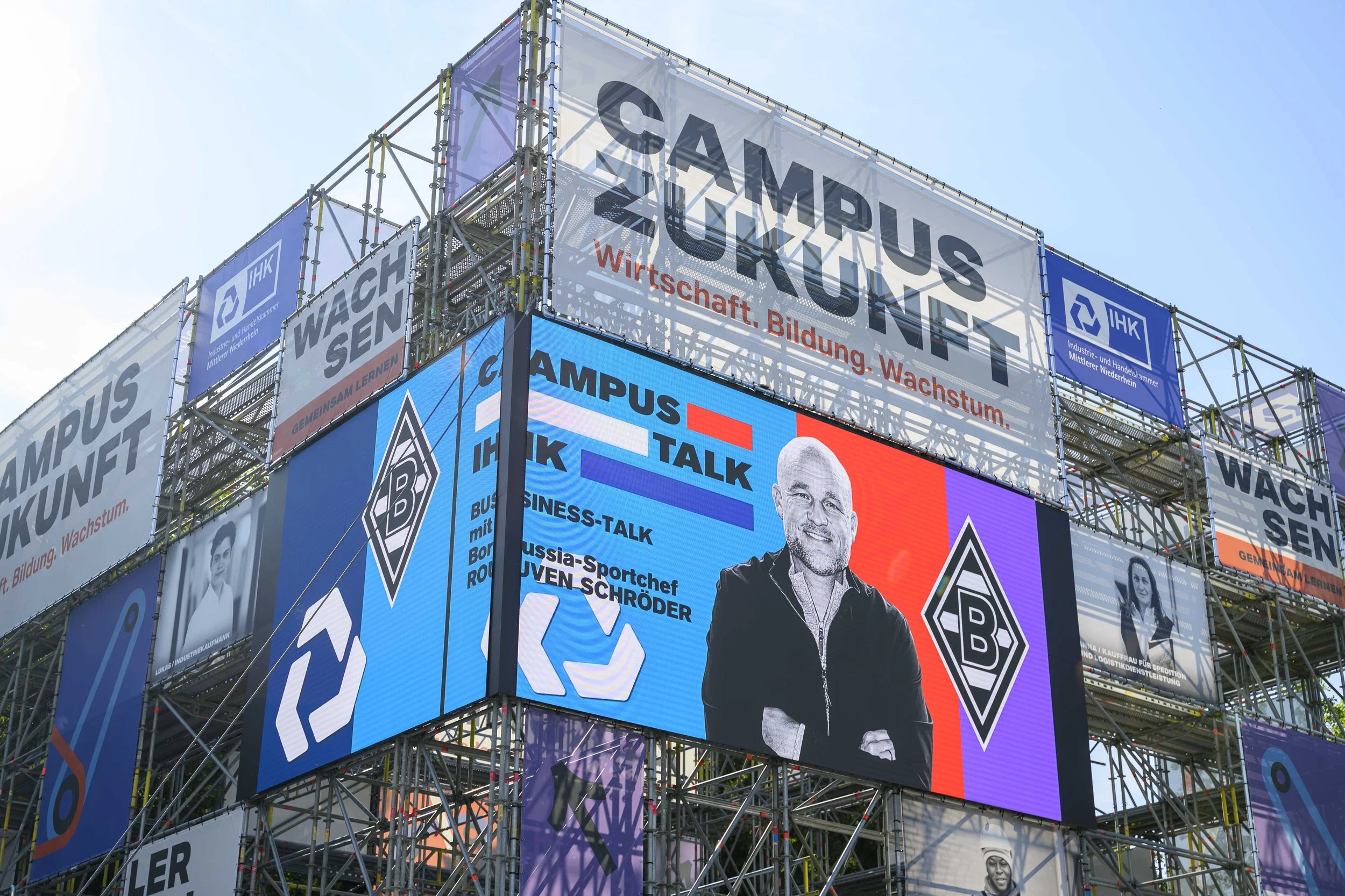 Mehrere große digitale Werbetafeln an einer Stadtkulisse, die Werbung für die Hochschule Bochum (HB) und andere Bildungsevents zeigen, mit einem blauen Himmel im Hintergrund.