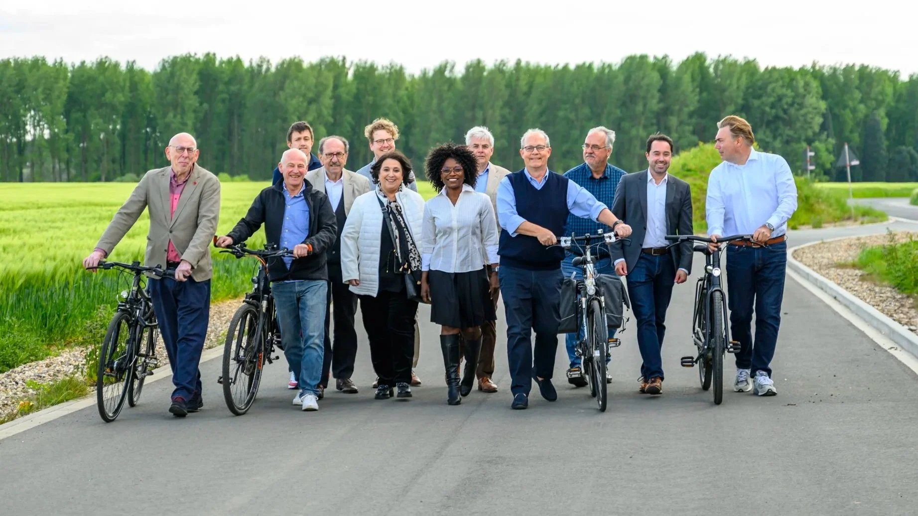 Eine Gruppe von Menschen, bestehend aus Männern und Frauen, die draußen auf einer Straße gehen. Zwei Männer haben Fahrräder, alle lachen und unterhalten sich, umgeben von grünen Feldern und Bäumen.
