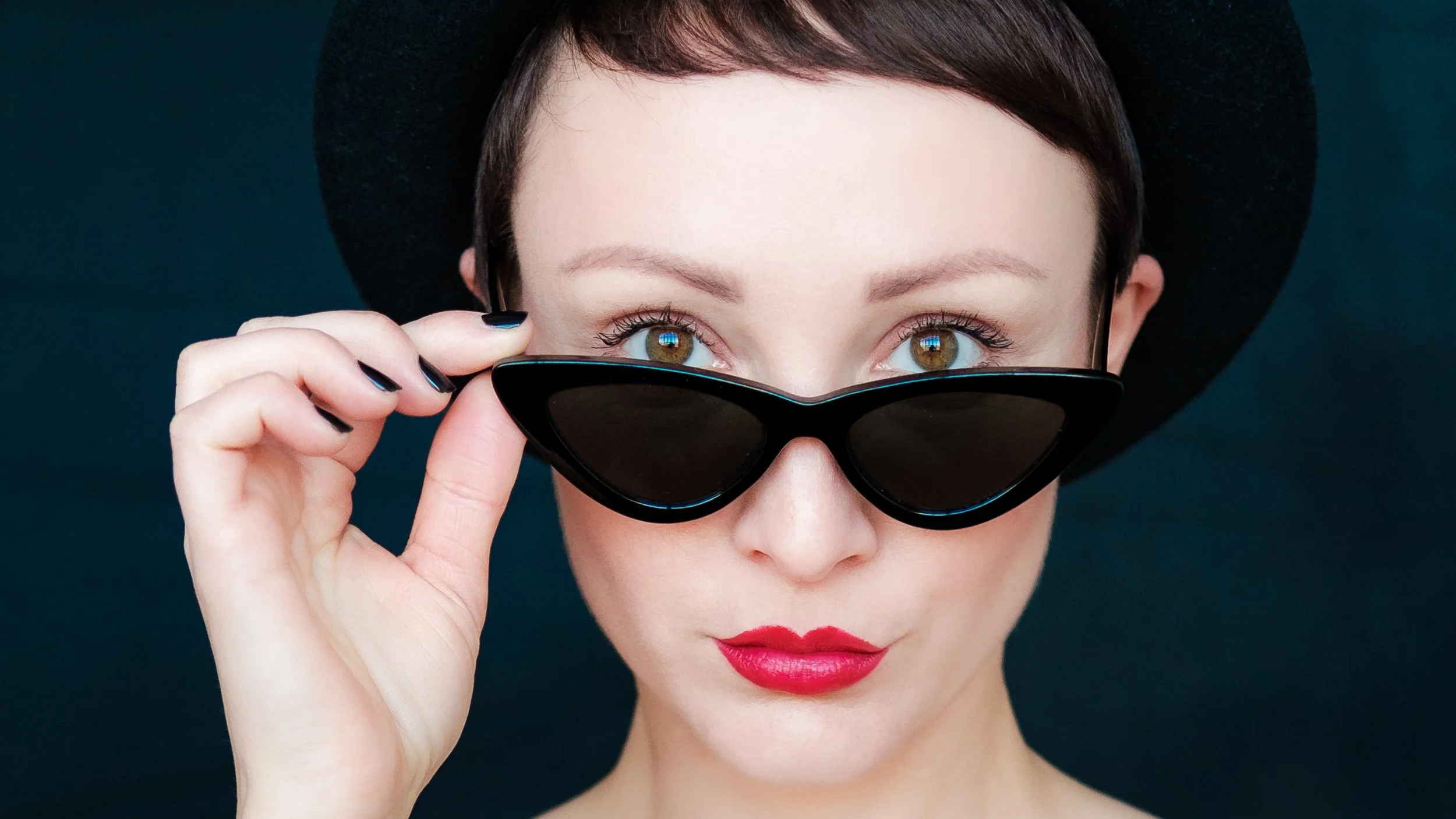 Junge Frau mit roten Lippen, who hat dunkle Haare und trägt eine schwarze Sonnenbrille und einen schwarzen Hut, schaut direkt in die Kamera.