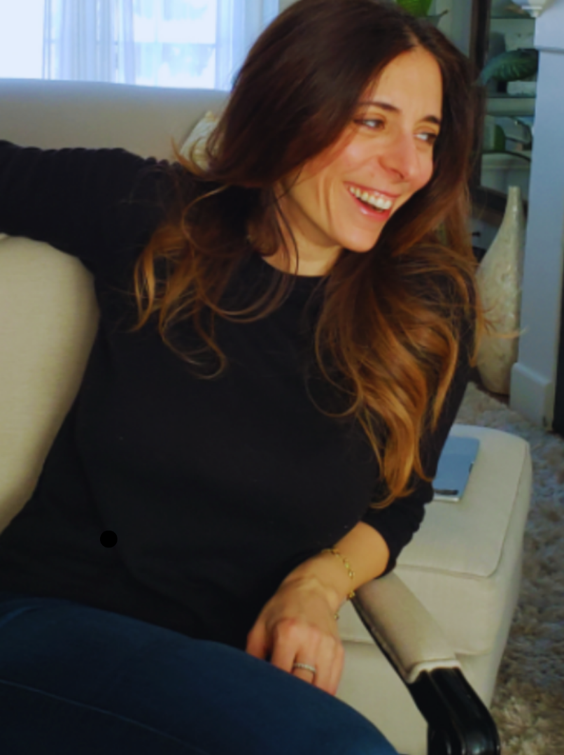 A woman with long brown hair sitting on a beige sofa, smiling and looking to her right, in a cozy living room with sunlight coming through a window in the background.