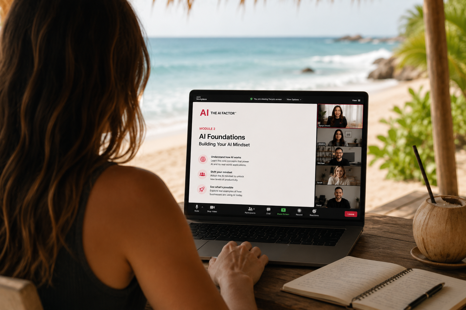 Woman with wavy hair sitting at a wooden table on a beach, participating in a virtual AI training session on a laptop, with a coconut drink nearby.