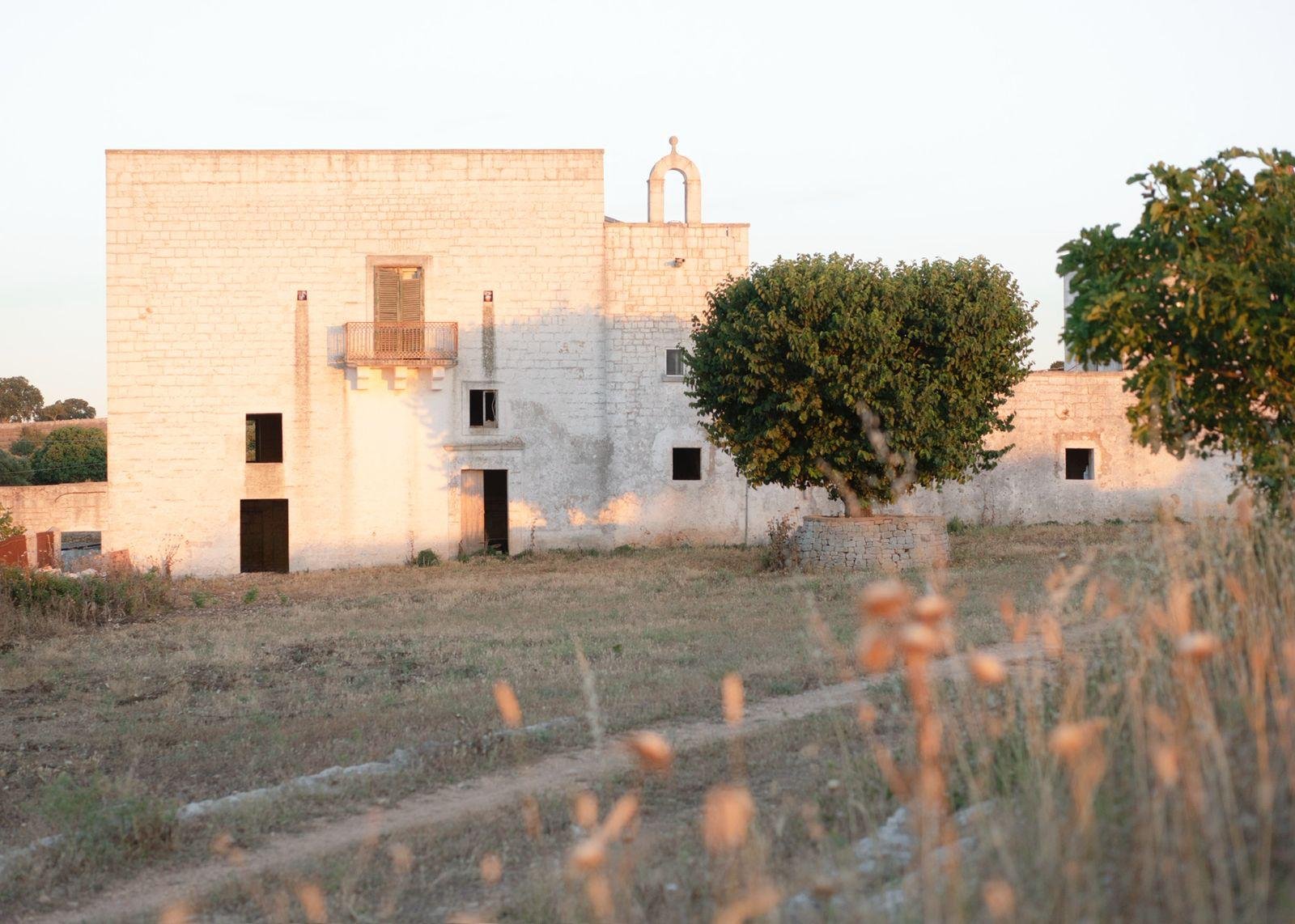An old stone building with multiple windows and doors, surrounded by dry grass and two green trees, under a clear sky, during sunset.