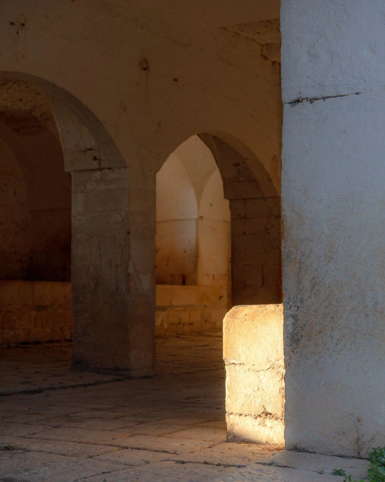 An old stone structure with arches and columns, illuminated by warm lighting, with a stone floor and textured walls.