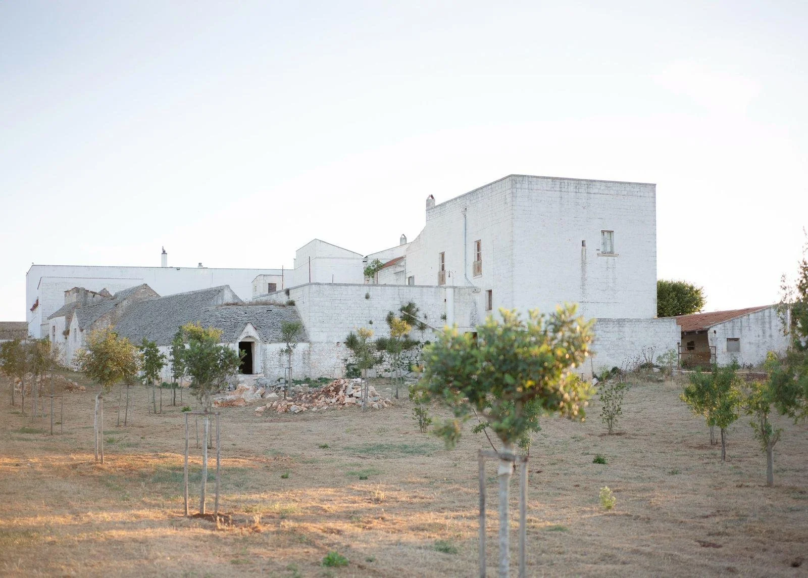 A rural scene with a cluster of white buildings, some with stone roofs, surrounded by small trees and dry grass.