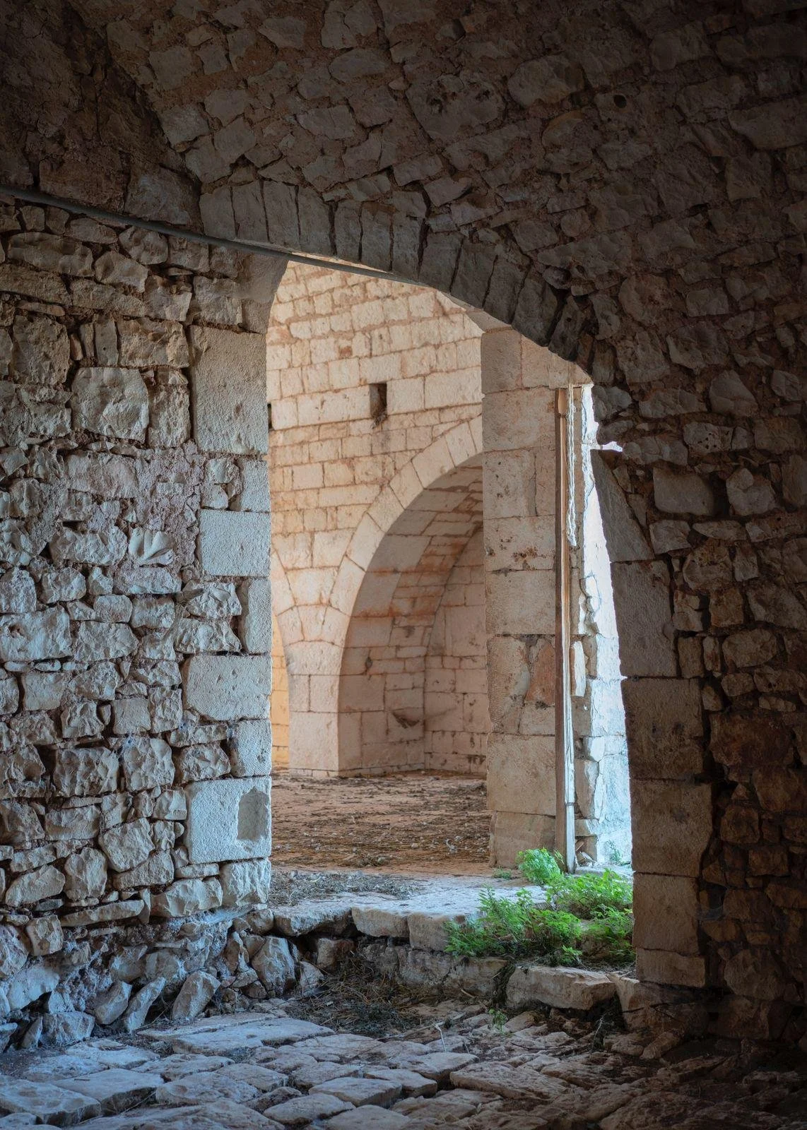 Stone archways and walls of an ancient structure, with sunlight coming through an opening, and some greenery on the ground.