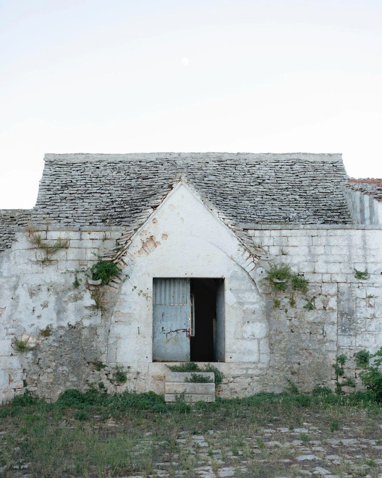 An old, weathered building with a sloped roof made of stone tiles, a small arched entrance with a metal door, and overgrown vegetation around the entrance area, under a clear sky with the moon visible.