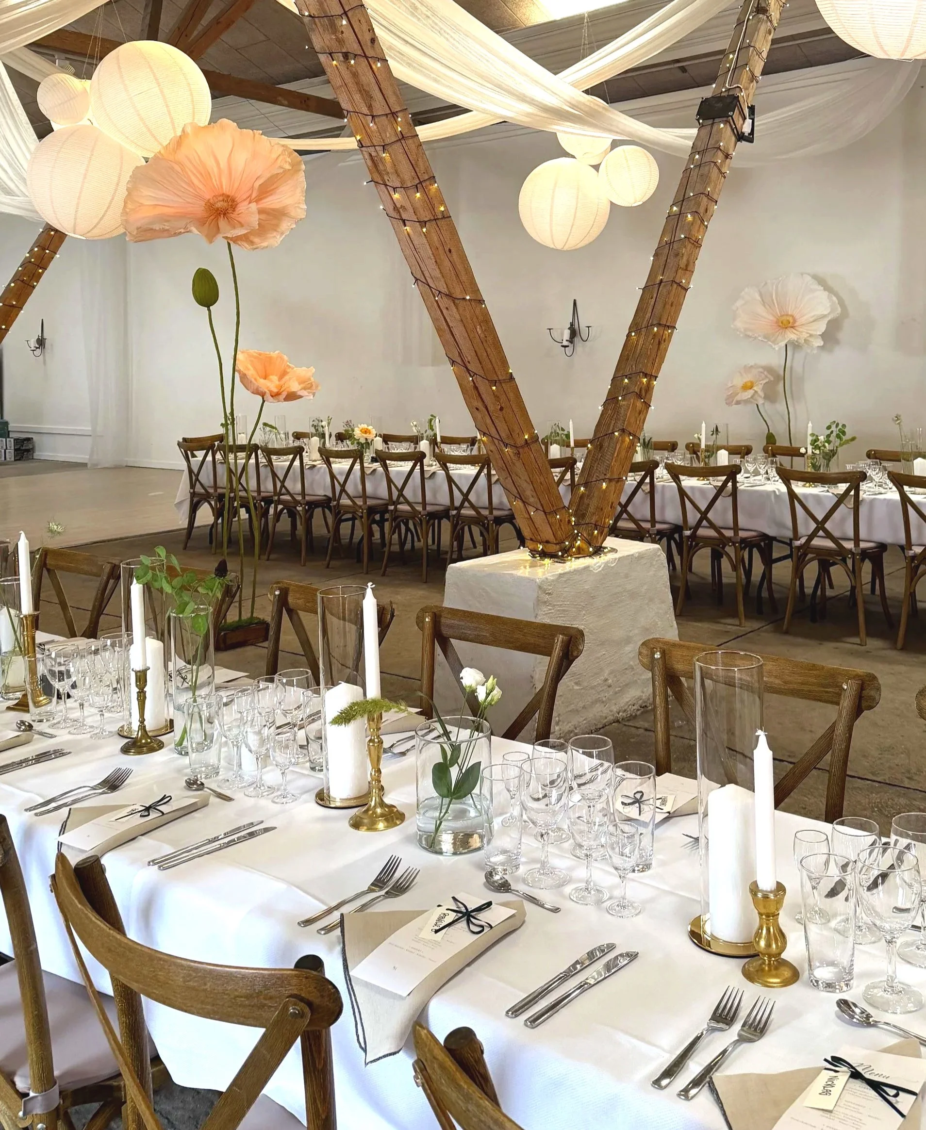 Elegant banquet table set with white tablecloth, gold candle holders, candles, glassware, menus, and small green plant arrangements. Decorated with large paper flowers and fairy lights, in a spacious room with draped fabric decorations and round pape