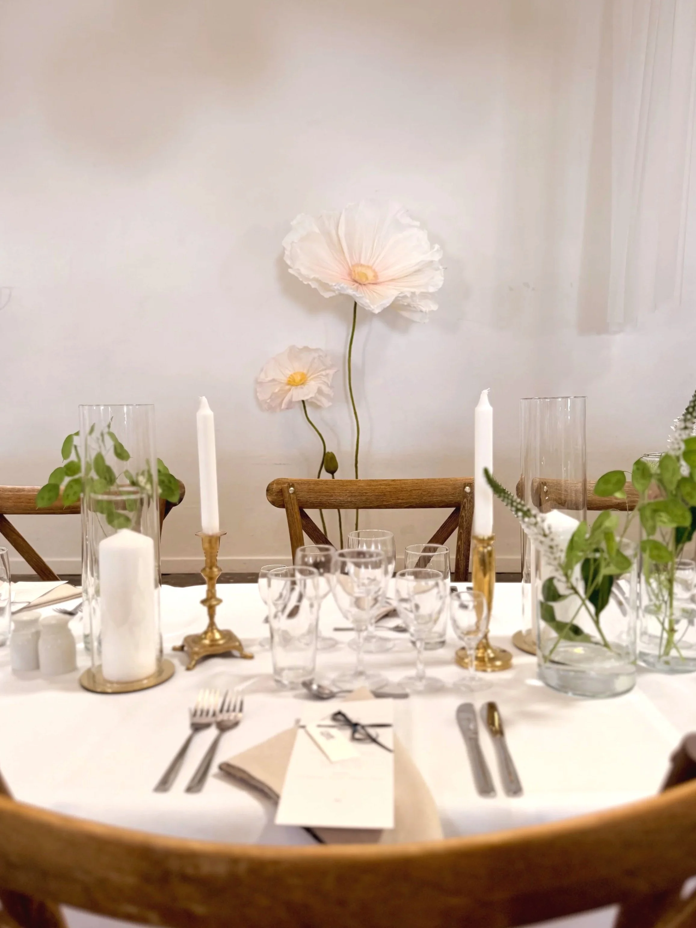 A dining table set for a formal event with white tablecloth, glassware, cutlery, candles, and greenery. In the background, two large white paper or fabric flowers on the wall, with a white wall and ceiling.