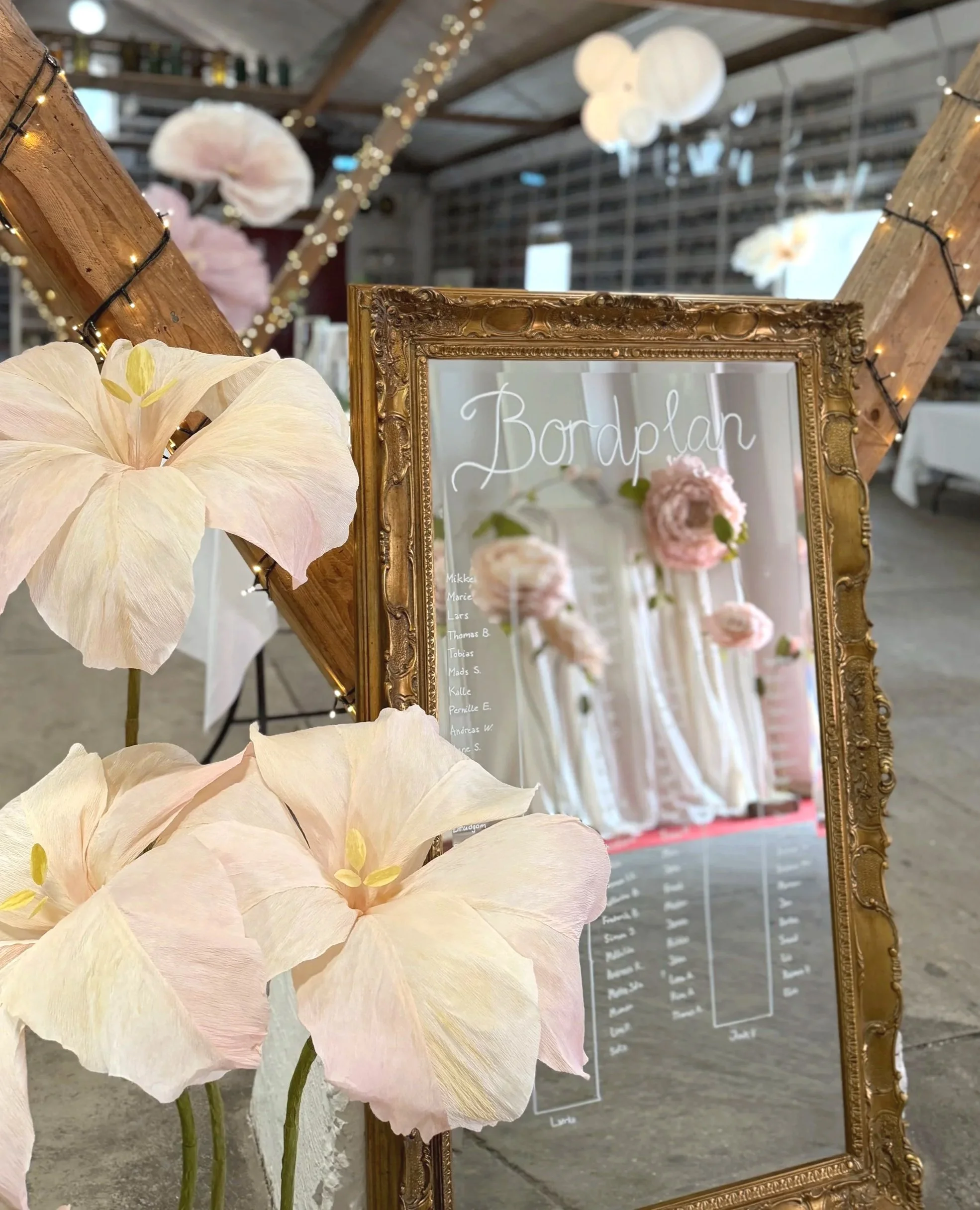 A decorated display with large light pink and cream-colored flowers, a gold ornate mirror, and a background showing a barn-like setting with draped fabric, fairy lights, and a table with clothing, possibly for a wedding or special event.