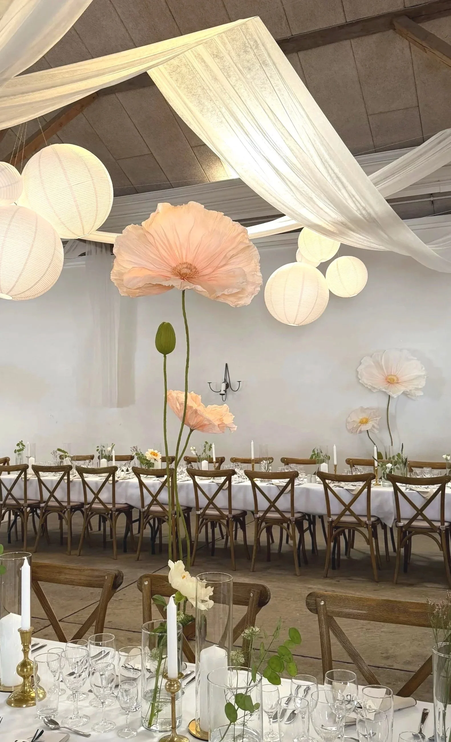 Elegant indoor event setup with long dining tables decorated with white tablecloths, glassware, and centerpieces, under hanging paper lanterns and fabric drapes, with large floral decorations resembling poppies.
