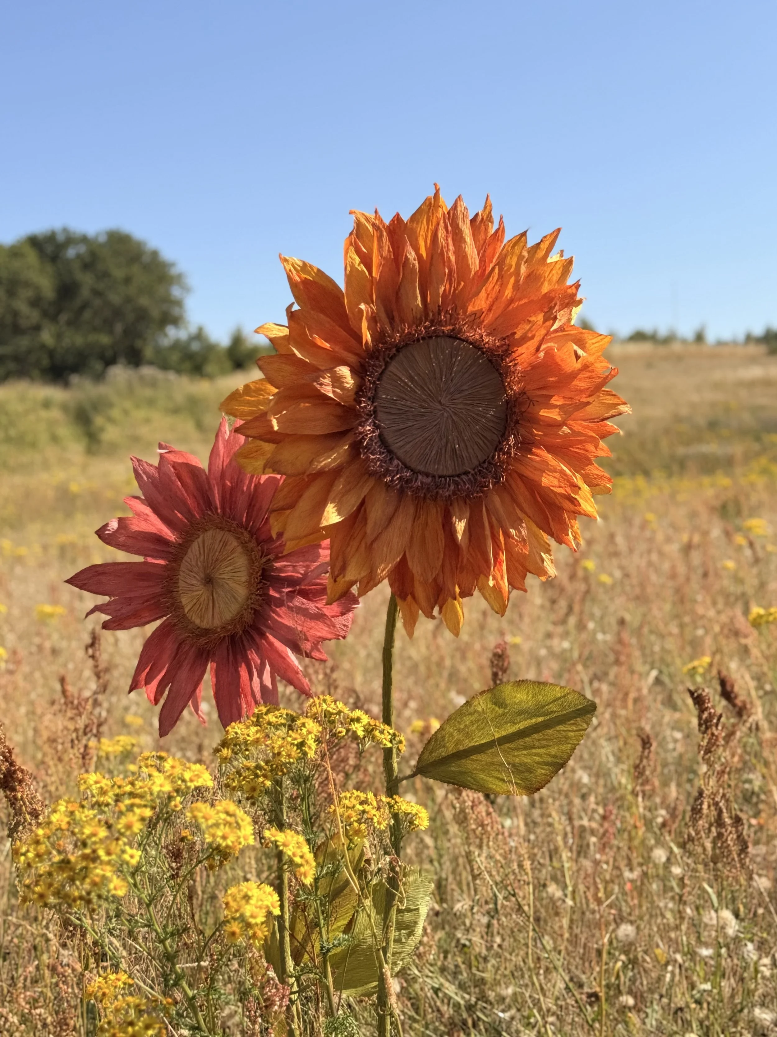 Close-up of a paper sunflower with vibrant orange petals and a dark center, standing tall in a field with yellow and pink flowers under a clear blue sky.