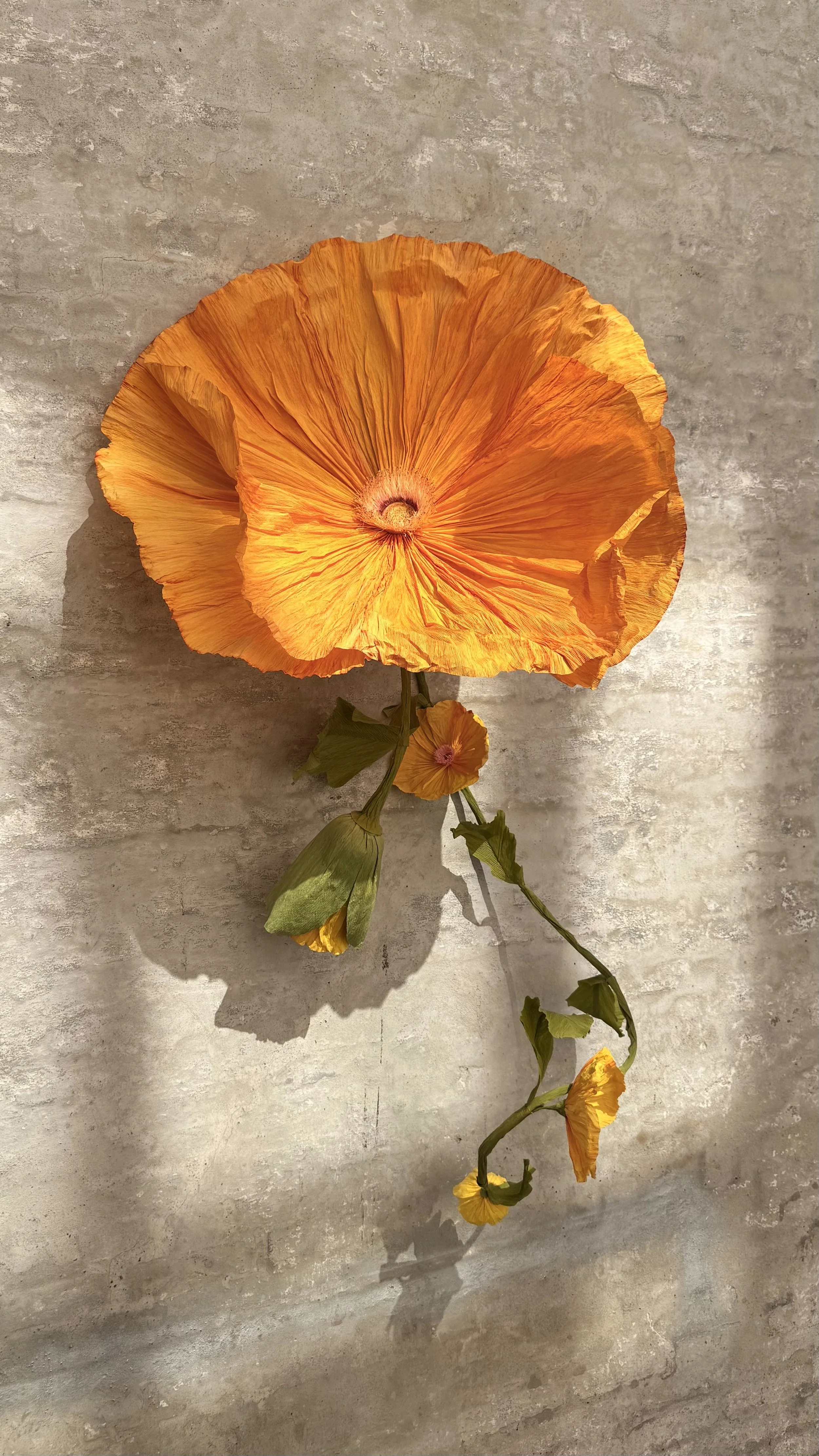 A large, vibrant orange artificial flower with layered, pleated petals and a central seed pod, attached to a twisted stem with secondary smaller flowers and green leaves, mounted on a textured concrete wall.