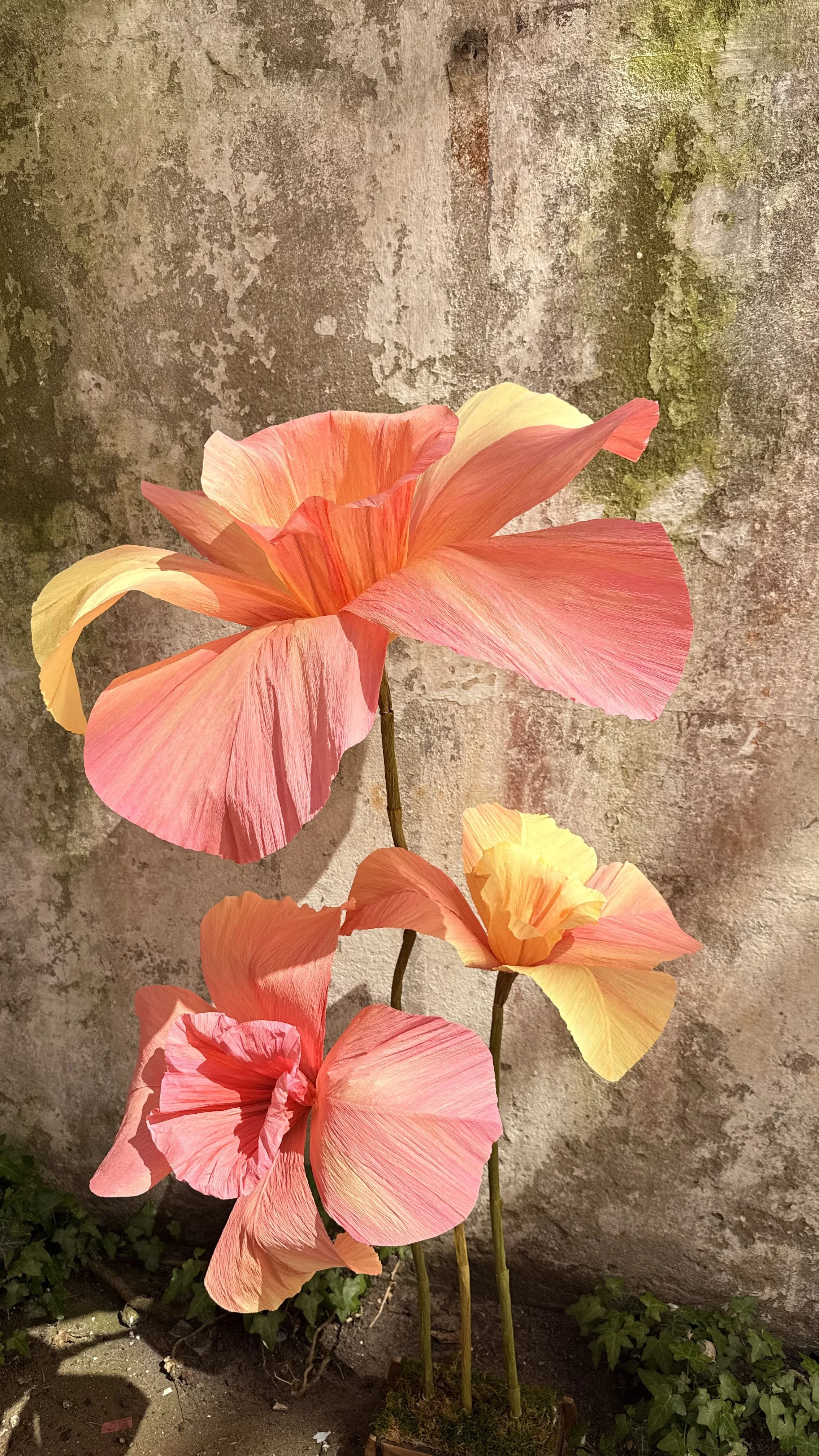 Artificial pink and yellow flowers in a garden against a weathered concrete wall.