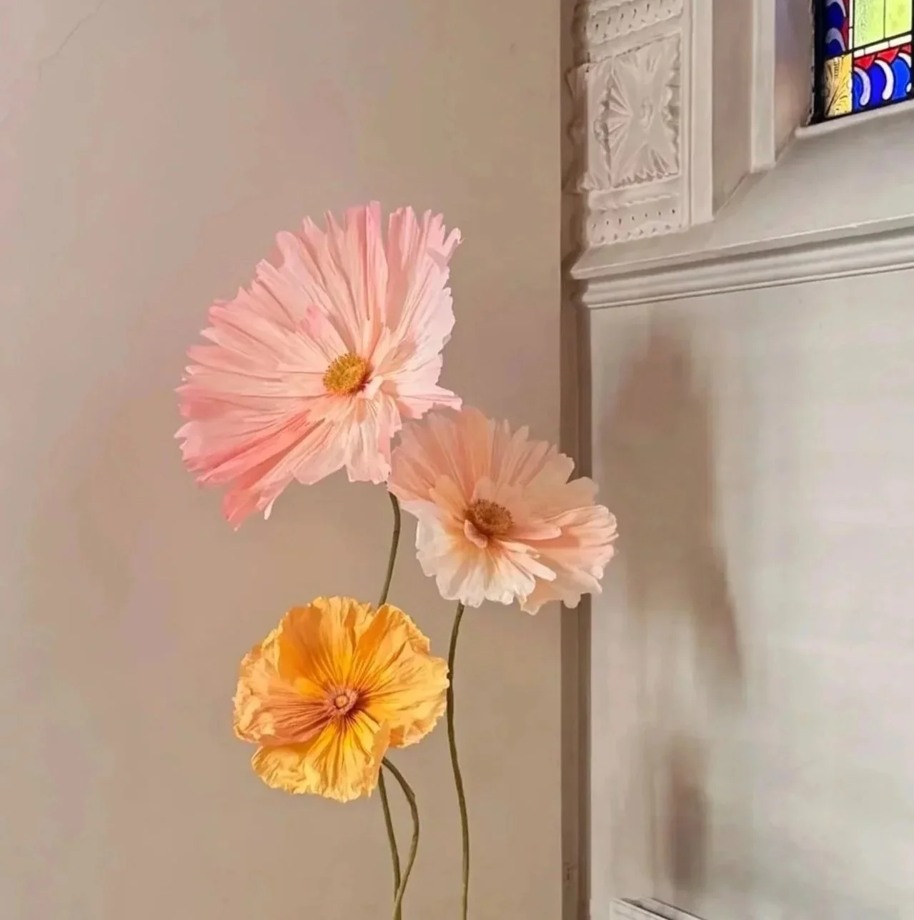 Three handcrafted giant paper flowers in pastel shades of pink, peach, and yellow with dark stems, set against an indoor background with a pale wall and a white piece of furniture