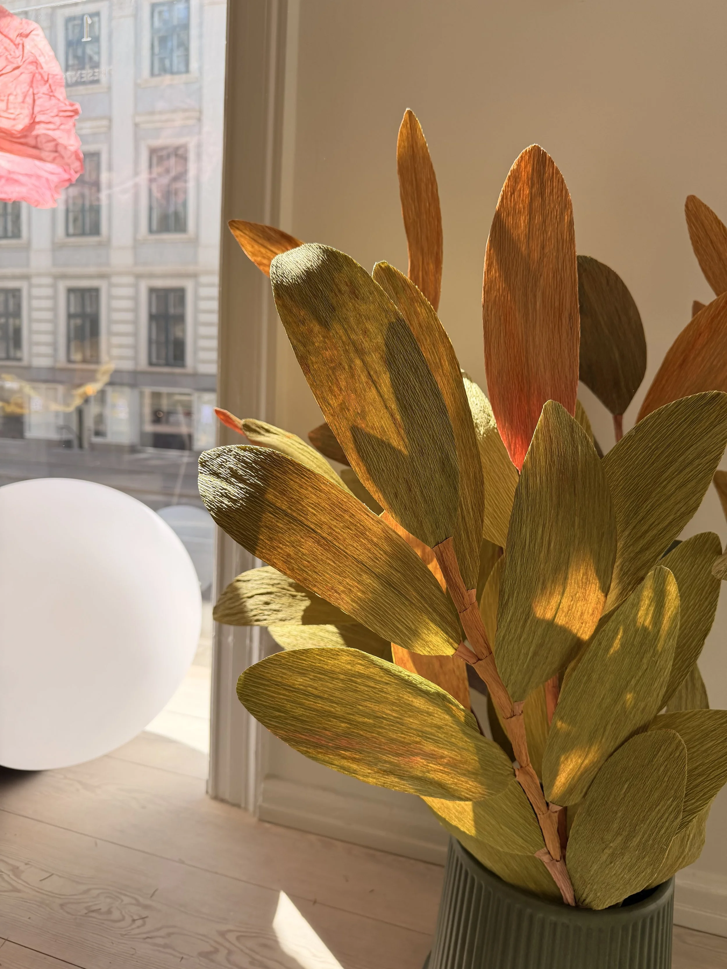 Artificial plant with large green and brown leaves in a gray pot on a wooden floor, sunlight shining through a nearby window.