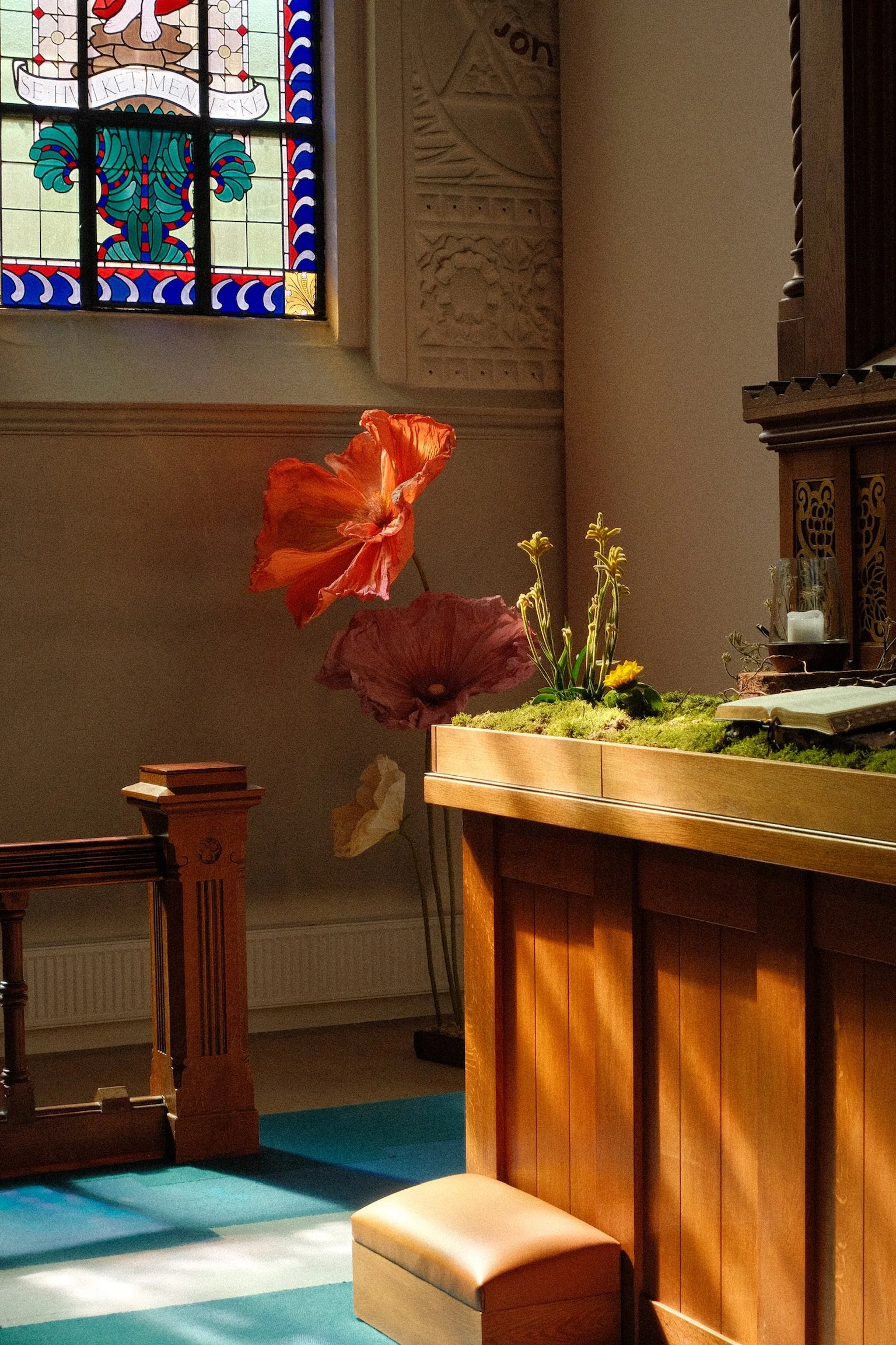 A church interior with a stained glass window, a carved stone wall, a wooden altar or pulpit, a small bench or stool, and a large flower arrangement with orange and pink flowers, green leaves, and yellow accents.