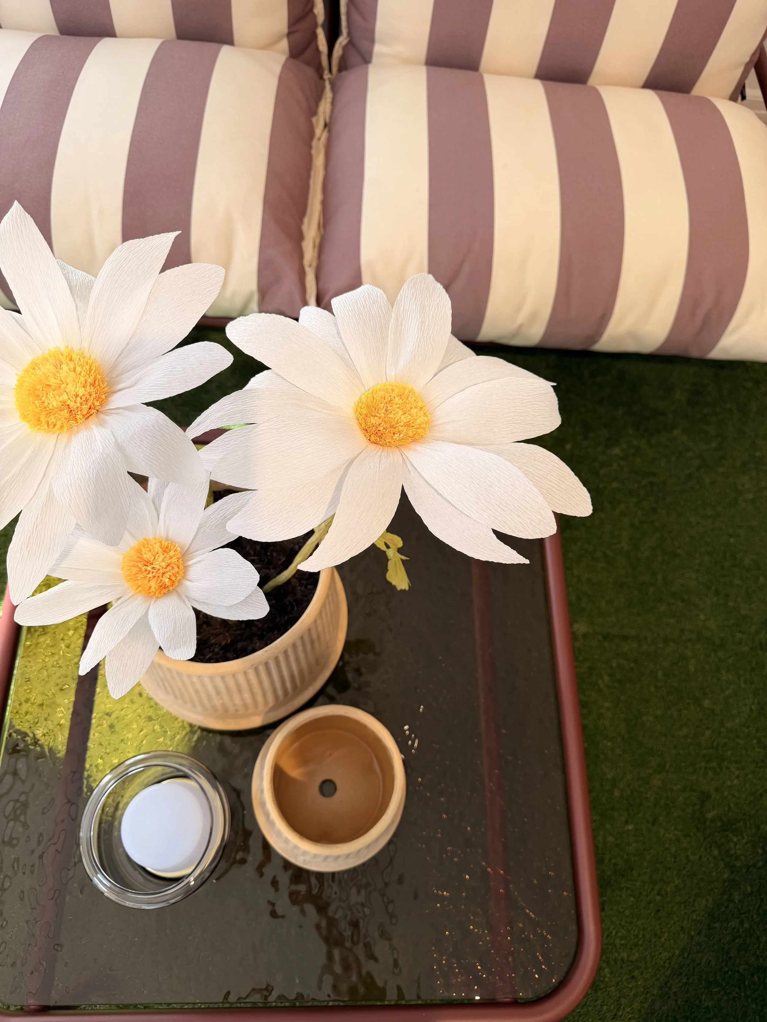Artificial daisies in a bowl on a wet black table with various small containers, some filled with liquids and one with white paint or cream, with a pink armrest in the background.