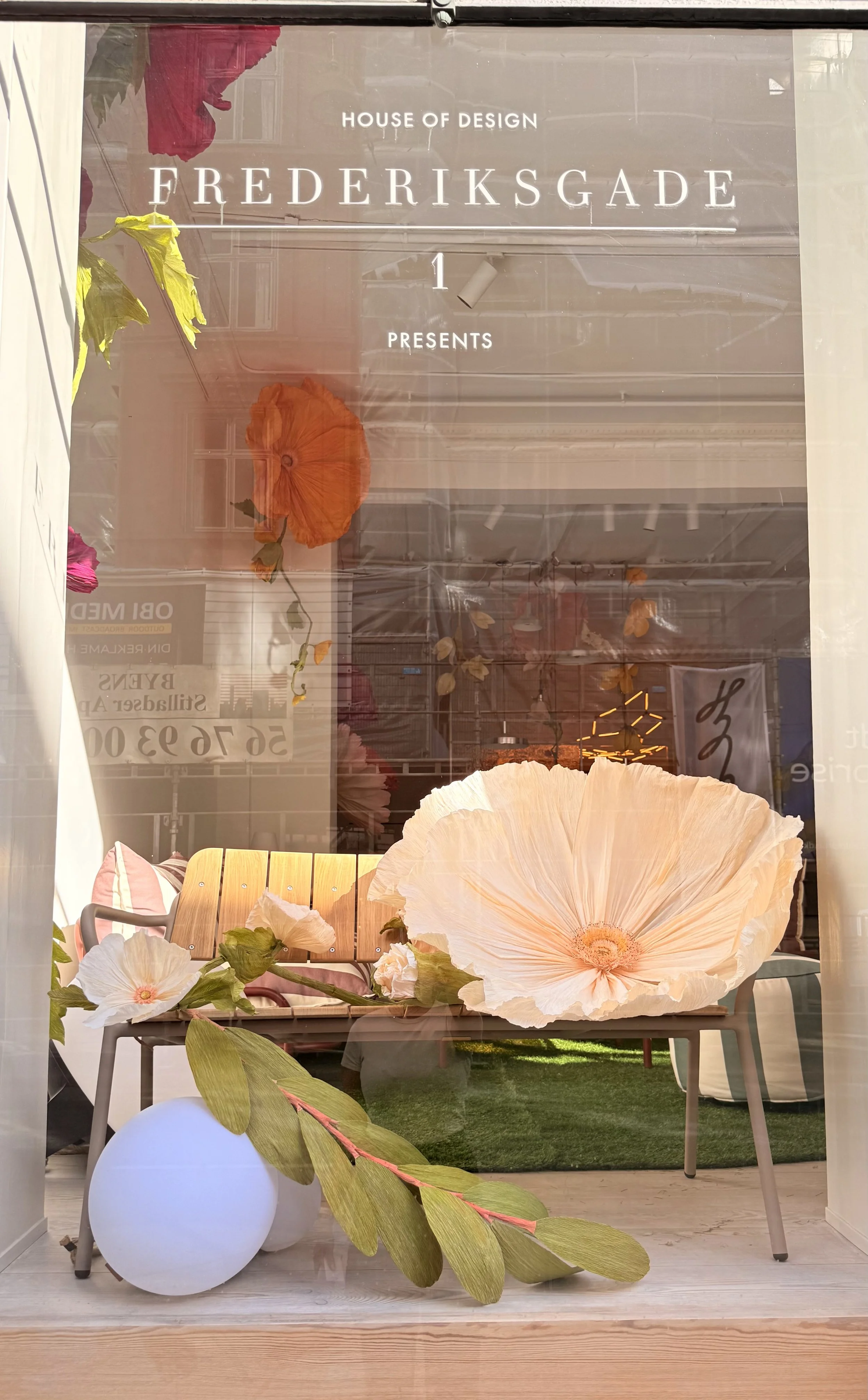 Store window display advertising a design house called Frederiks Gade at 1 Frederiks Gade, featuring large paper flowers, a glowing orb, and furniture inside the store