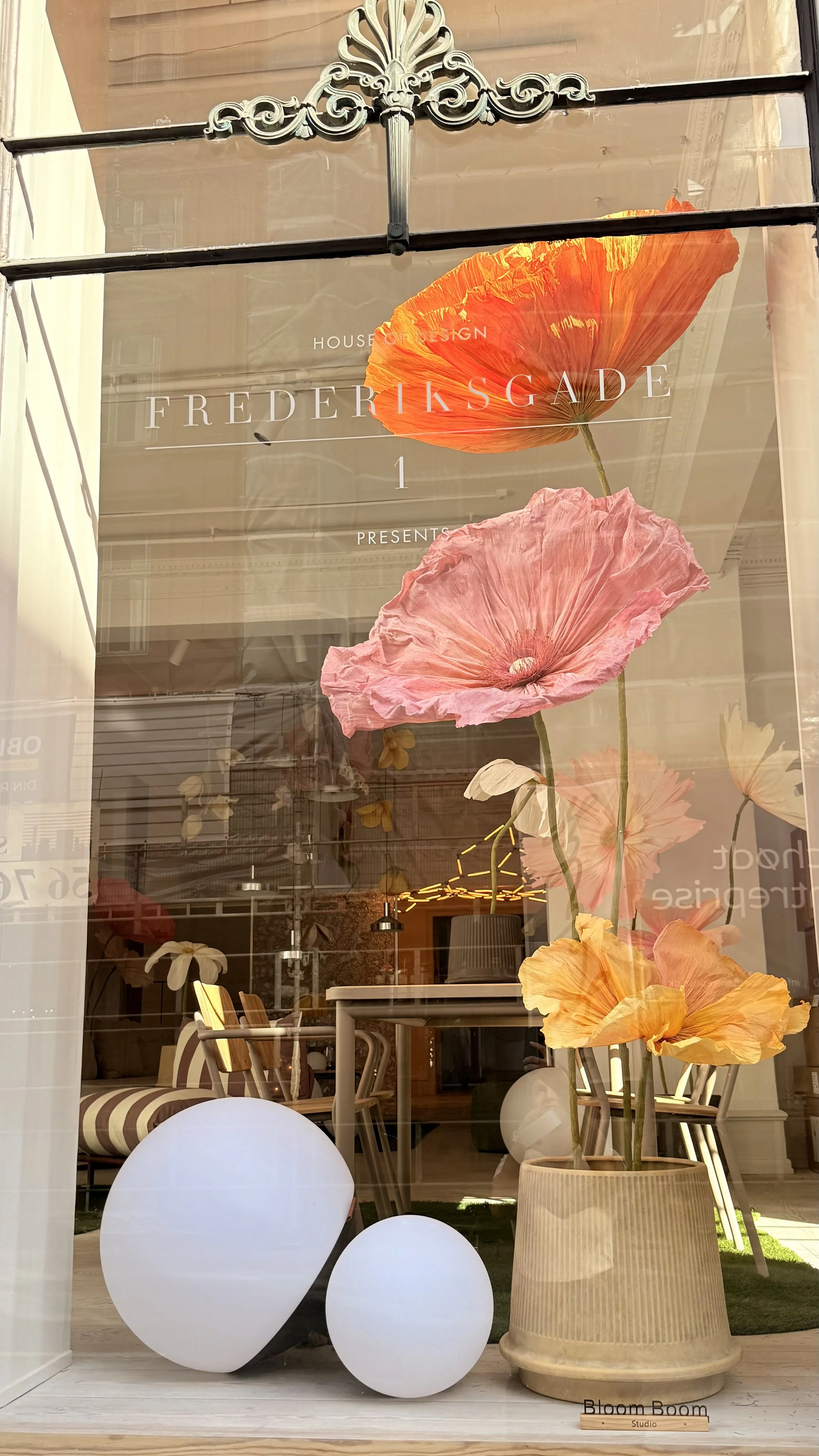 Display window with large pink and orange paper poppy flowers, white spherical decor, and a beige ribbed planter with a small wooden sign reading 'Bloom Boom Studio.' Inside, there are chairs with striped upholstery, a table, and hanging lights.