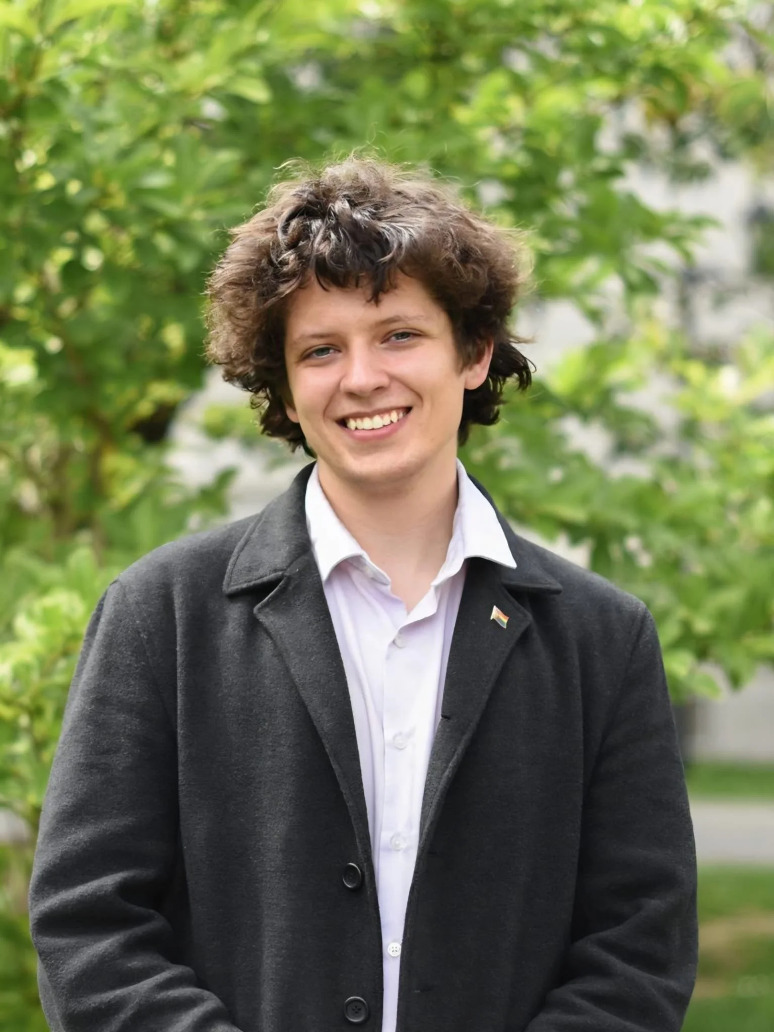 A young man with curly brown hair, wearing a gray coat over a white shirt, stands outdoors with green foliage in the background and a small rainbow flag pin on his lapel, smiling at the camera.