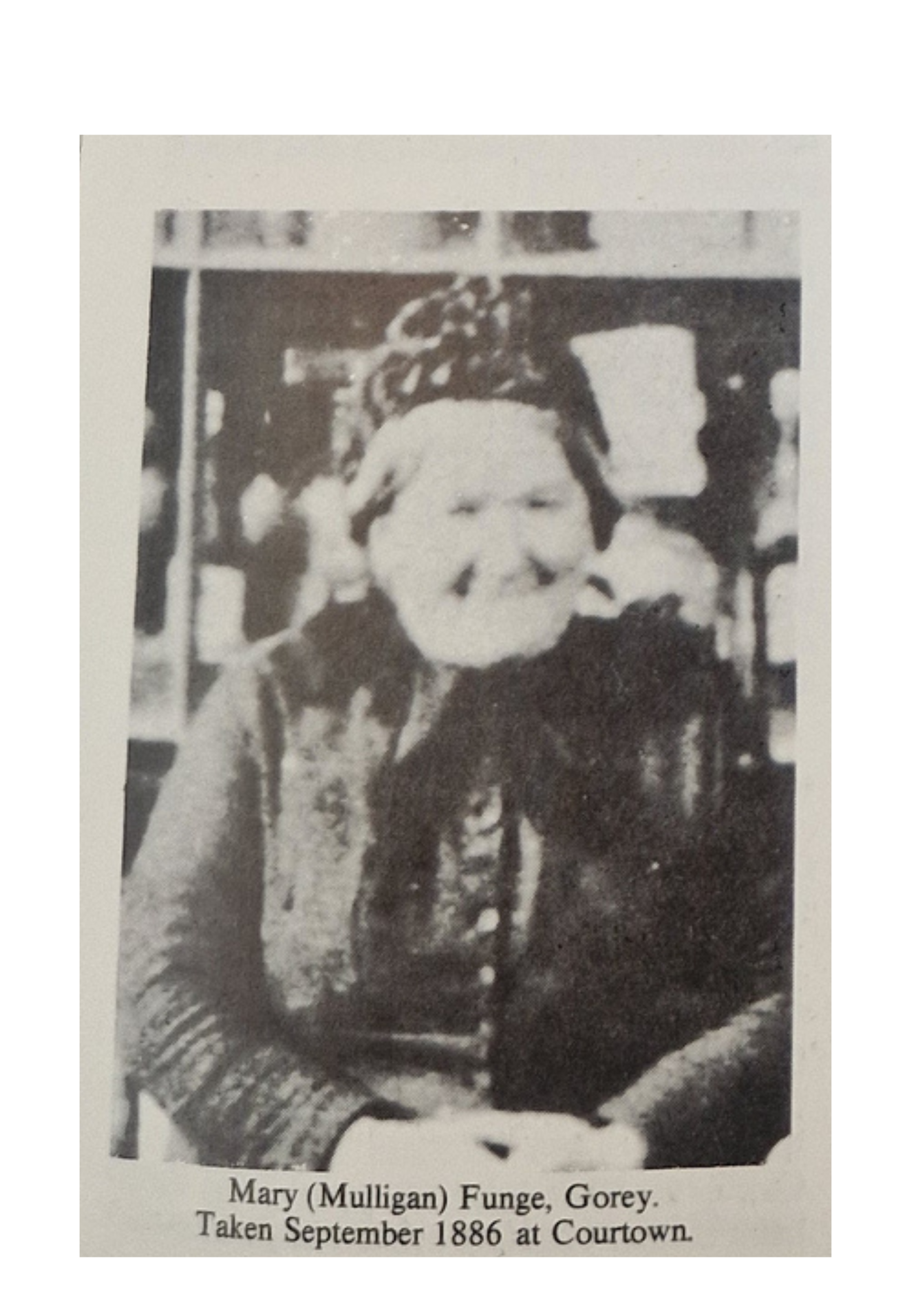 Black and white historical photograph of a woman named Mary (Mulligan) Funge, Gorey, taken in September 1886 at Courtown. She is dressed in late 19th-century attire, standing with her hands on her hips, in front of a building with a window.