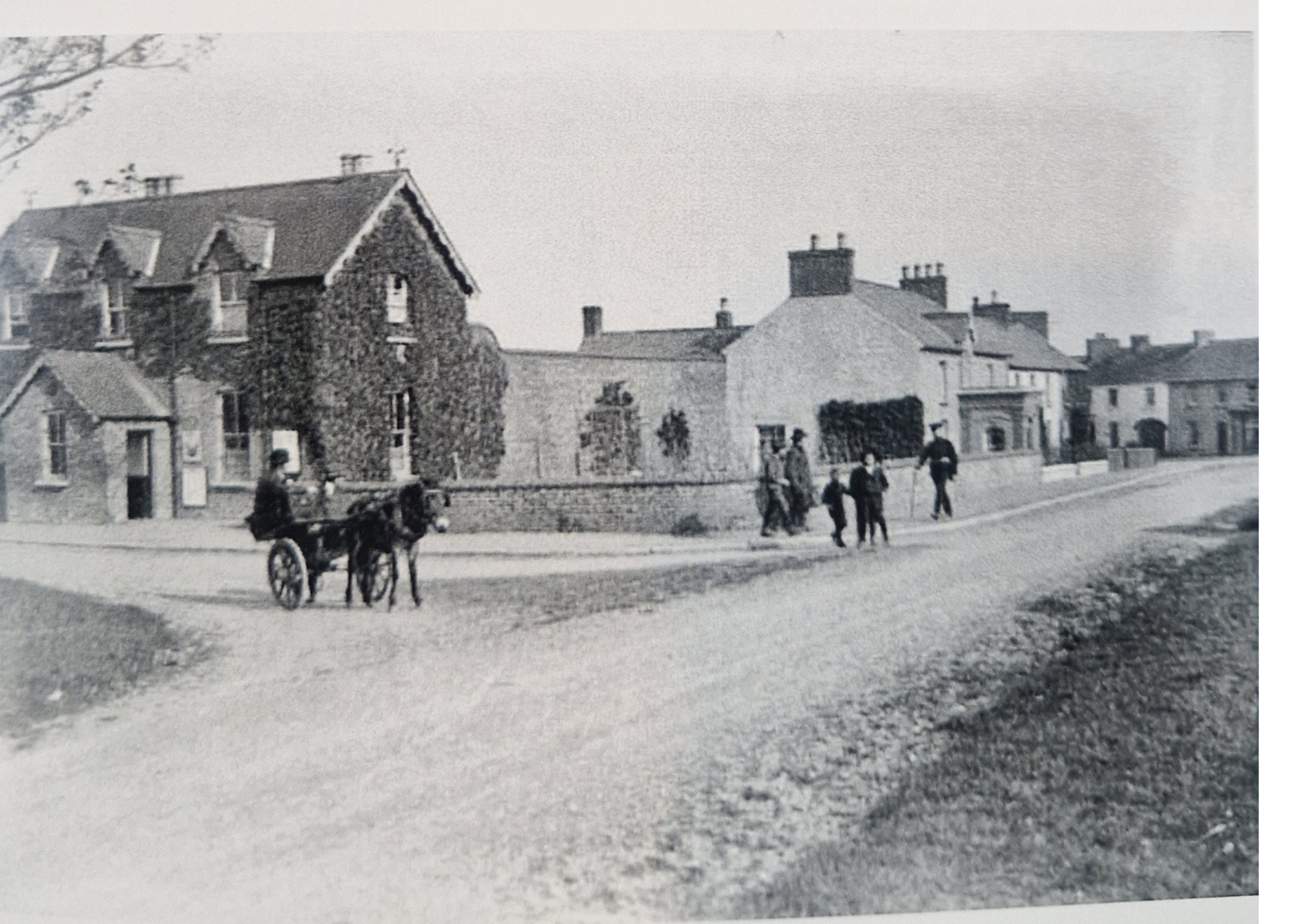 Black and white vintage photograph of a residential neighborhood with several houses, some with chimneys, and a group of children and a man standing on the street, some on bicycles and one on a cart pulled by a horse.