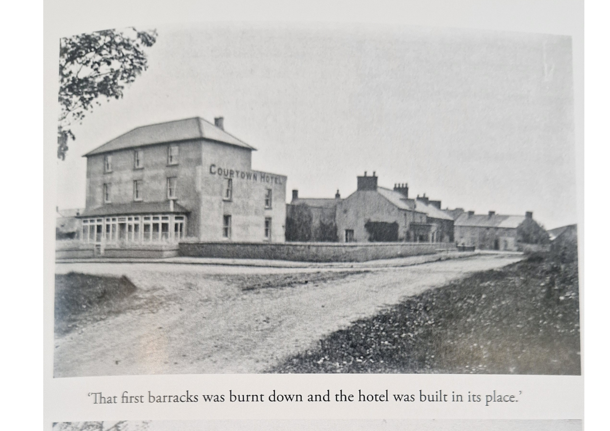 Black and white photograph of a hotel building named 'County Town Hotel', with an inscription visible on the building. The area in front of the hotel looks undeveloped or under construction, with dirt and gravel. There are smaller buildings with chim