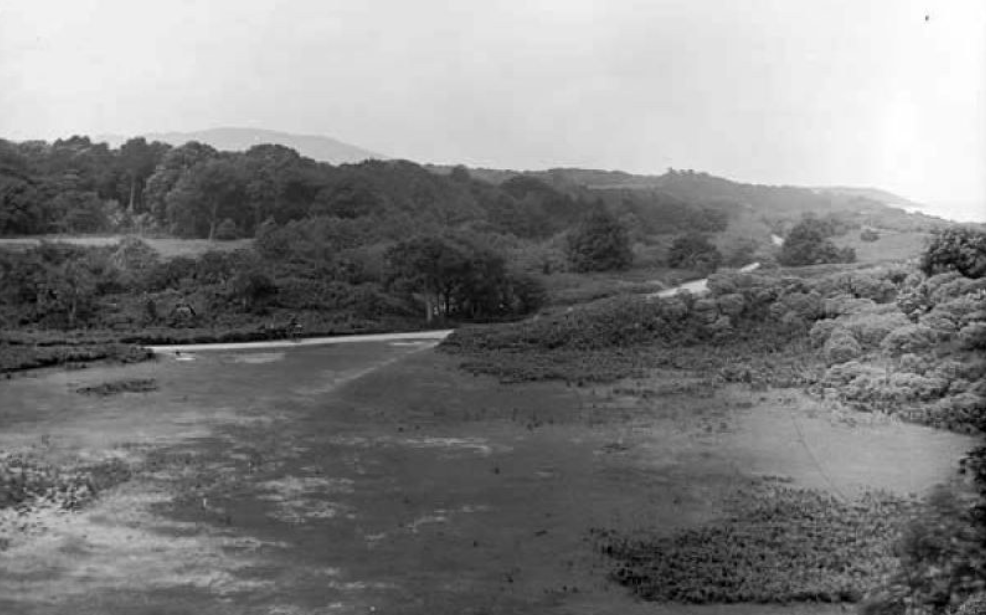 Black and white landscape photo of a river flowing through a rural area with trees and hills in the background.