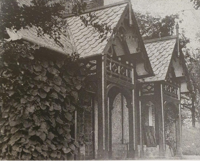 Black and white photograph of a rustic wooden house with a steep, shingled roof and decorative woodwork, surrounded by trees.