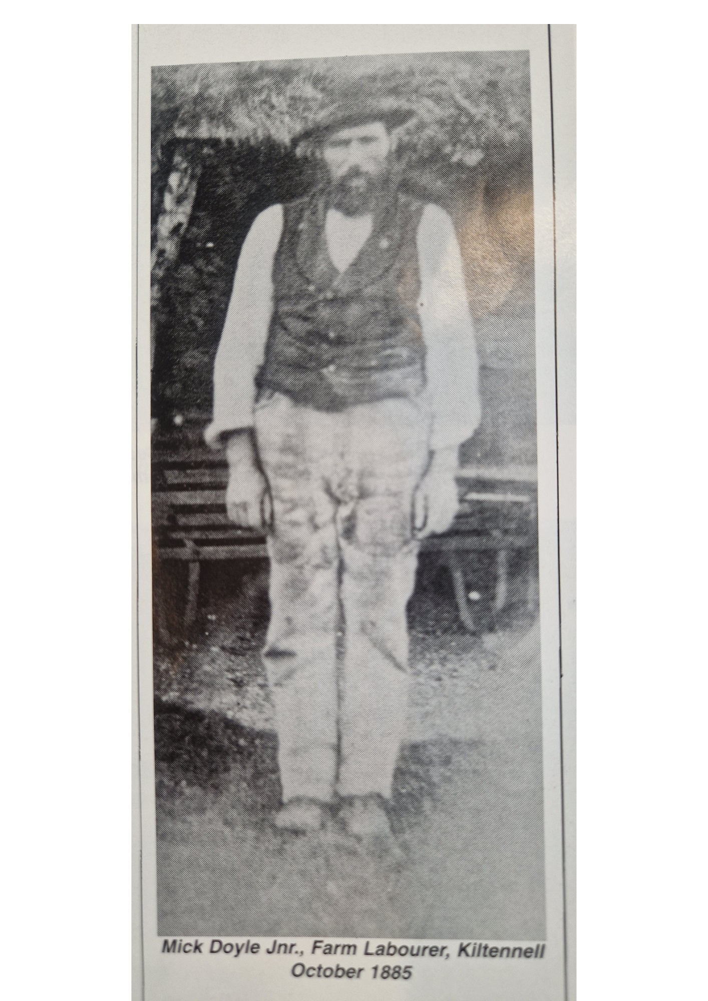 A black-and-white photograph of Mick Doyle Jr., a farm labourer from Kiltennell, standing outdoors with trees in the background, wearing traditional rural attire including a vest, shirt, and trousers, holding a stick, dated October 1885.
