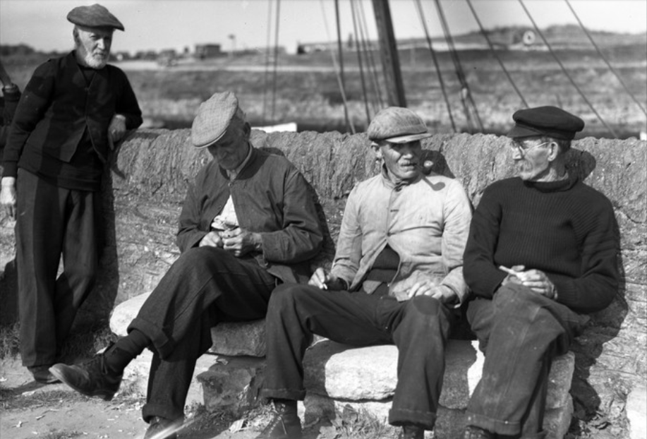 Four men sitting and standing outdoors, sitting on a stone wall, engaged in conversation. The background shows wire fencing and a rural landscape.