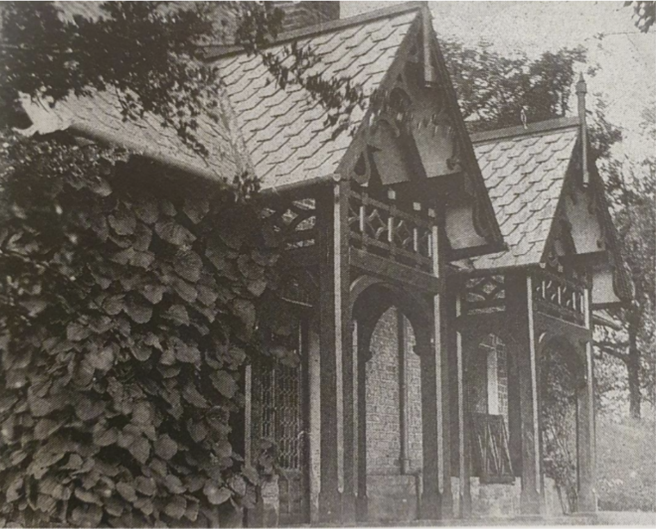 A black and white photograph of a decorated wooden structure with pointed roofs, arched openings, and a small balcony, surrounded by trees and foliage.