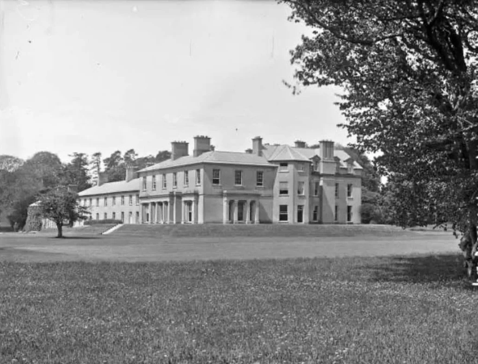 Black and white photo of a large, historic mansion with multiple stories, chimneys, and a front porch, surrounded by trees and a grassy lawn.