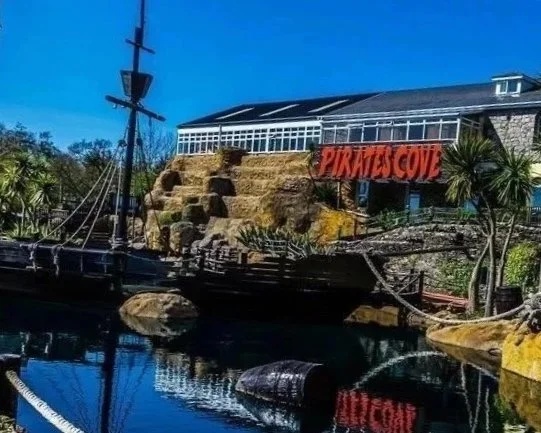 Pirates Cove sign with a pirate ship replica in front of a building, rocks, and water at a maritime-themed attraction.