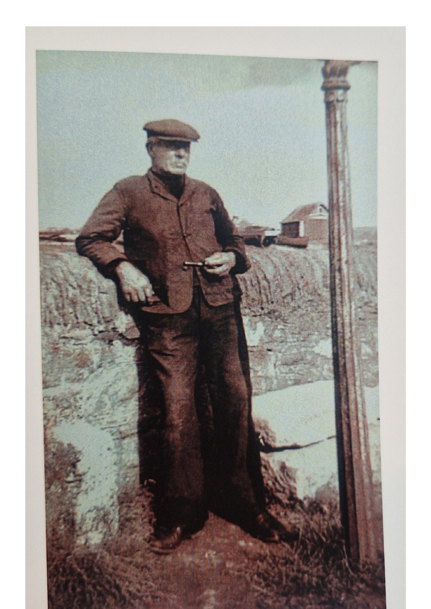 A black-and-white photo of a man standing outdoors in a rural setting, holding a cigarette, with a wooden pole or post next to him and small buildings in the background.